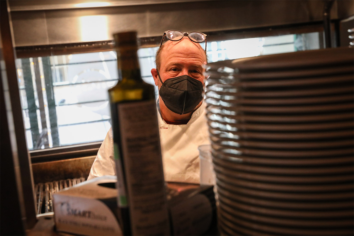 Frank Bonanno is seen from behind a stack of plates and a bottle of olive oil. He is in the kitchen of his restaurant, French 75.