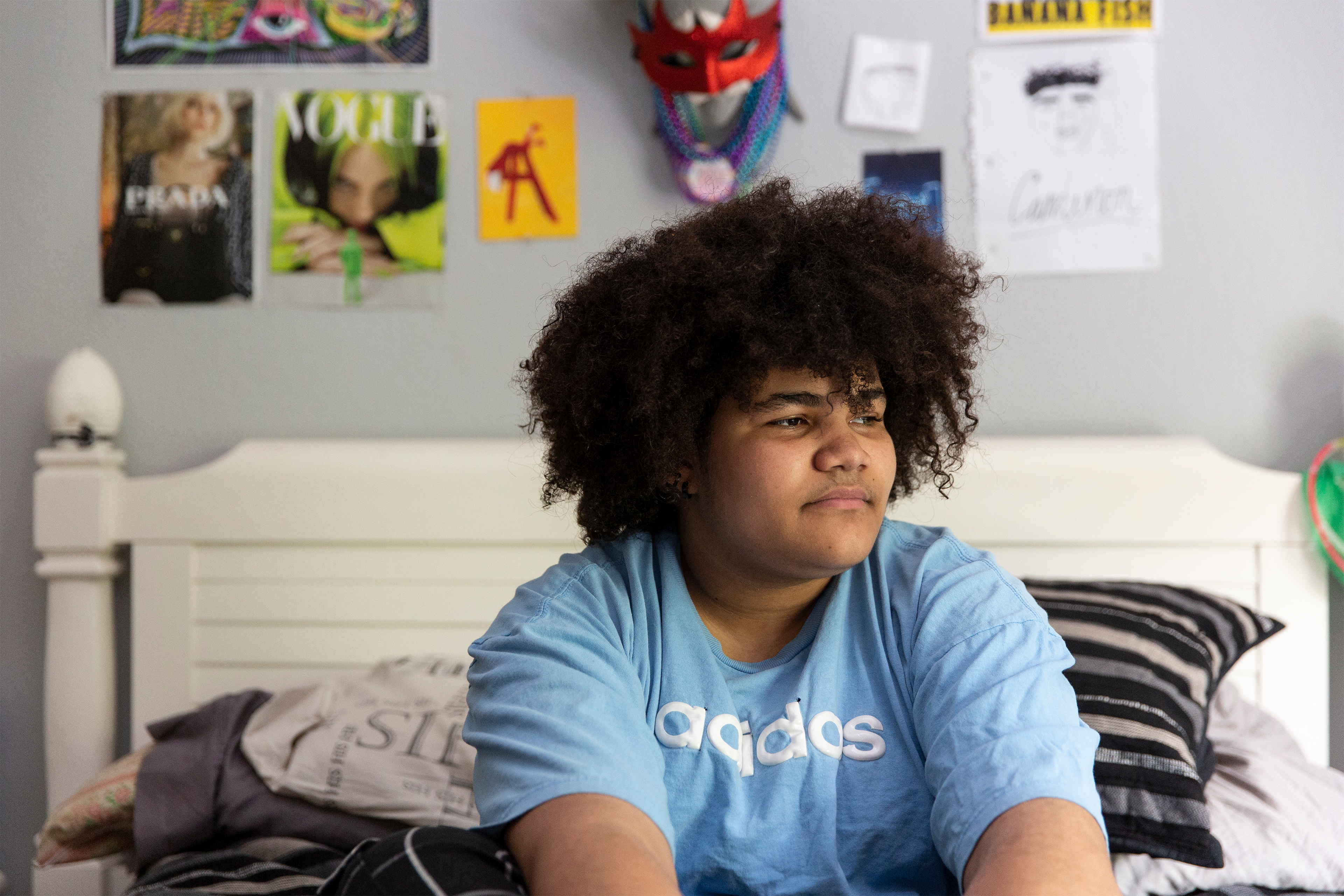 Cameron Wright sits on his bed at home, looking to the right. Posters and magazines decorate the wall behind him.