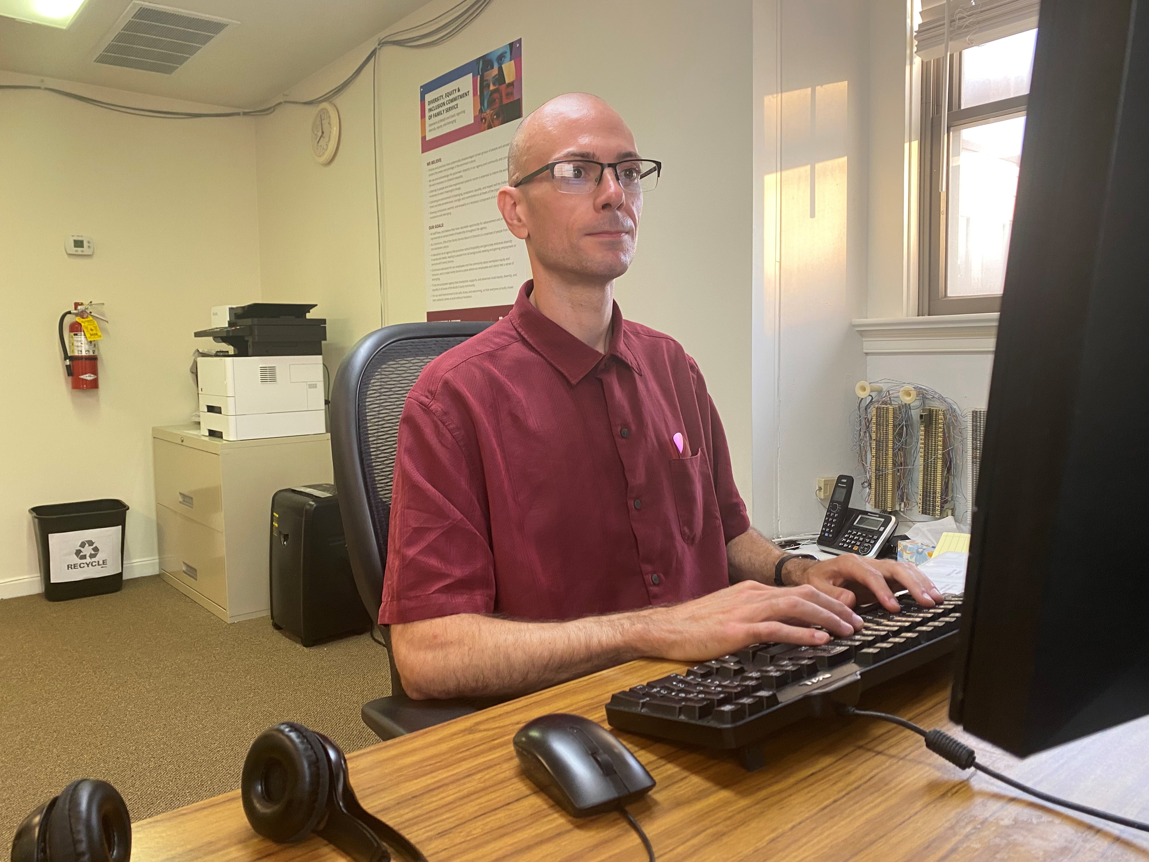 Michael Colluccio works at his desk. He wears a red shirt and rectangular glasses.