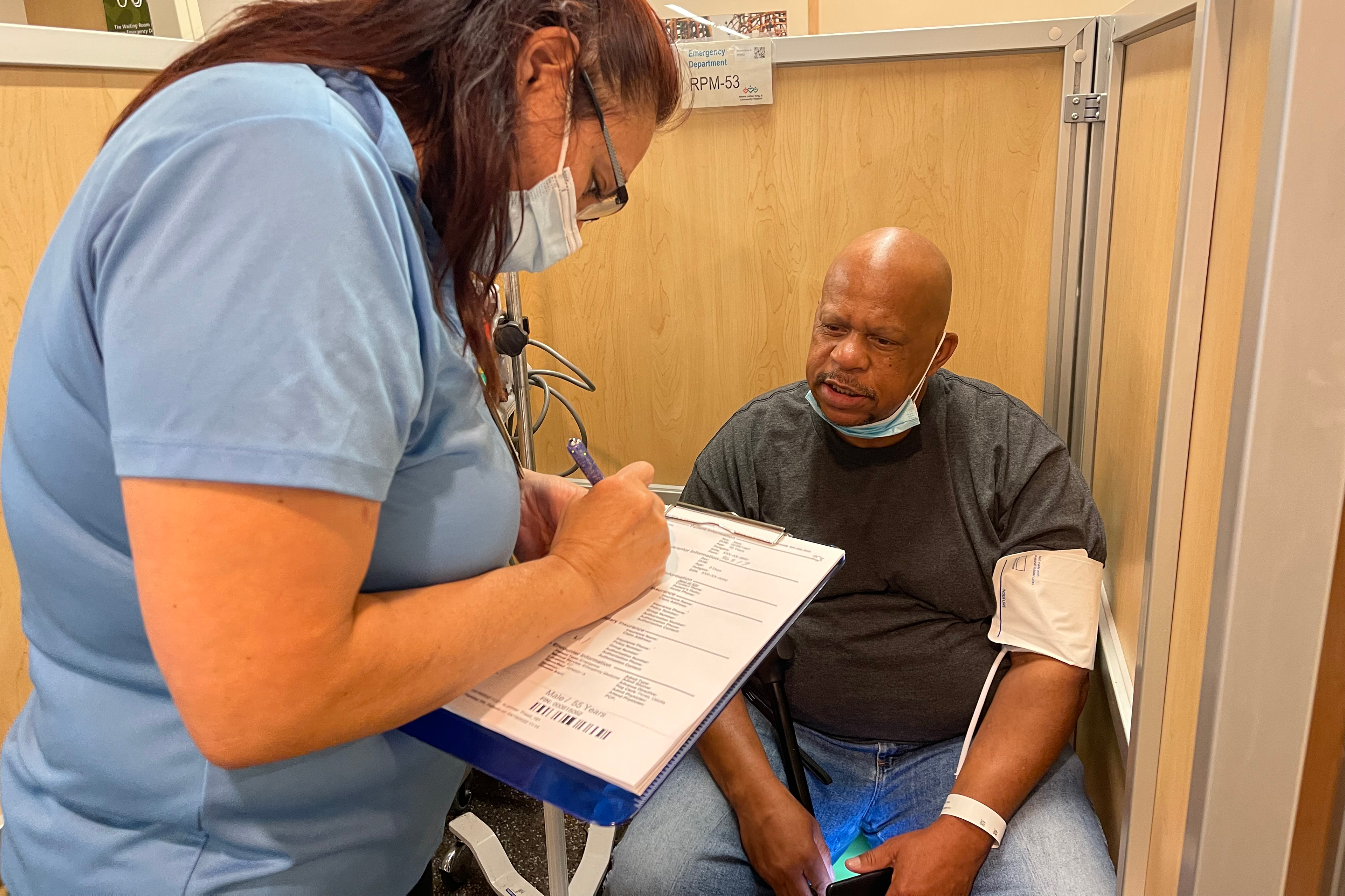 A photo shows Michael Reed sitting down in a hospital with a blood pressure cuff around his arm. A nurse is standing in front of him, writing on a clipboard.