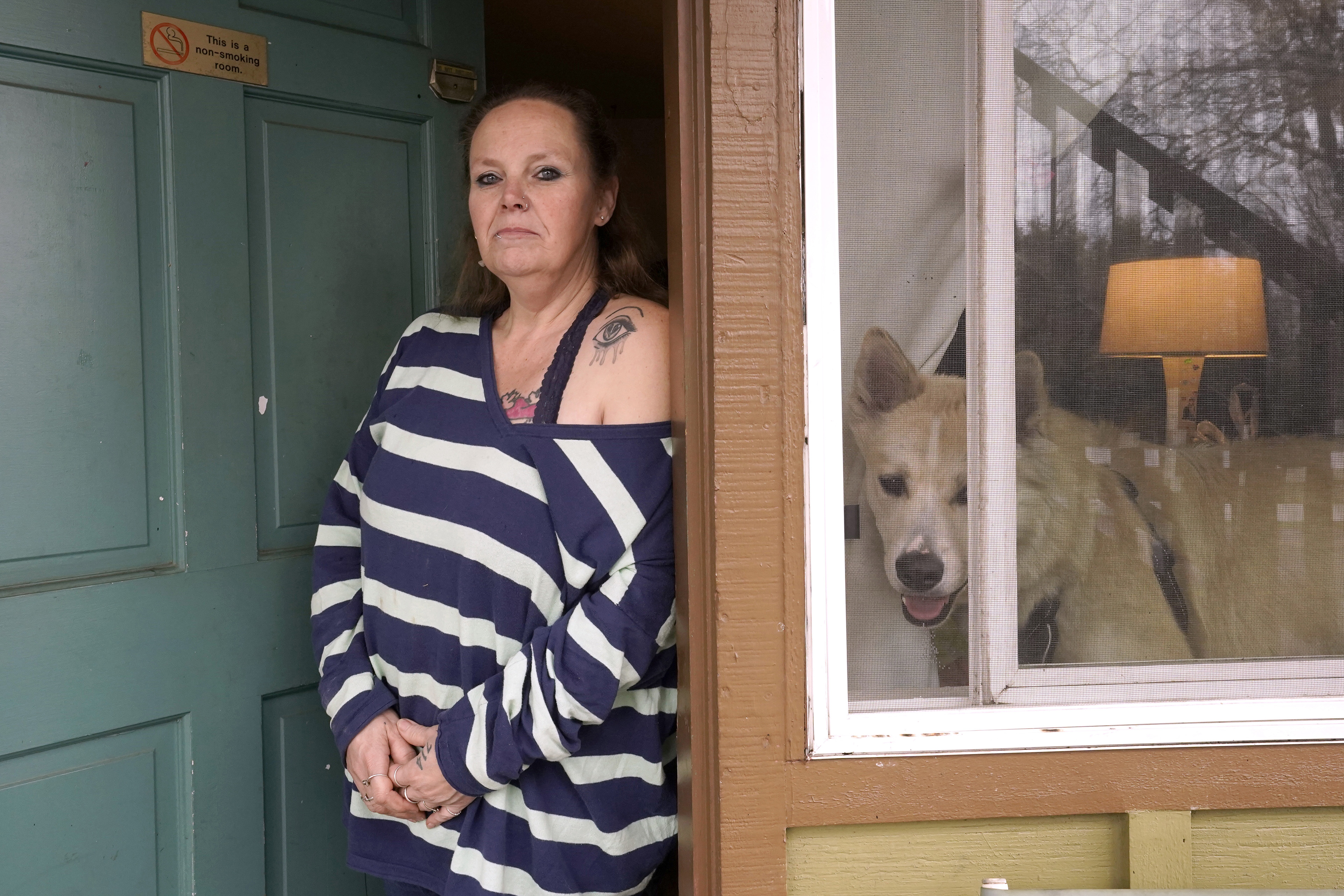Stephanie Lammers stands in the front door of her house and looks directly towards the camera. Her dog is visible in the window beside her.