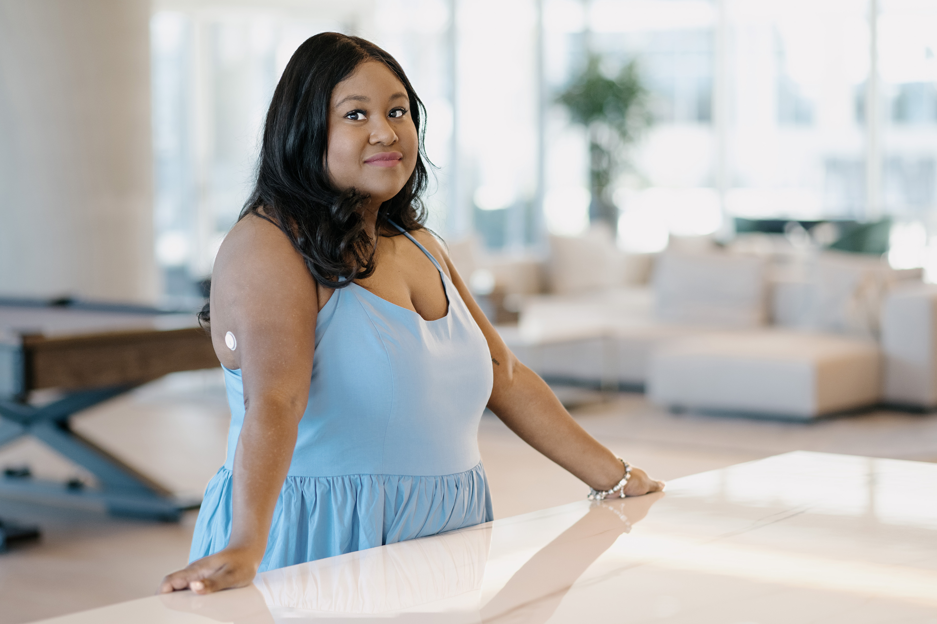 Mila Clarke stands in her home. She wears a pale blue dress and looks towards the camera. A couch and houseplant are faintly visible in the background.