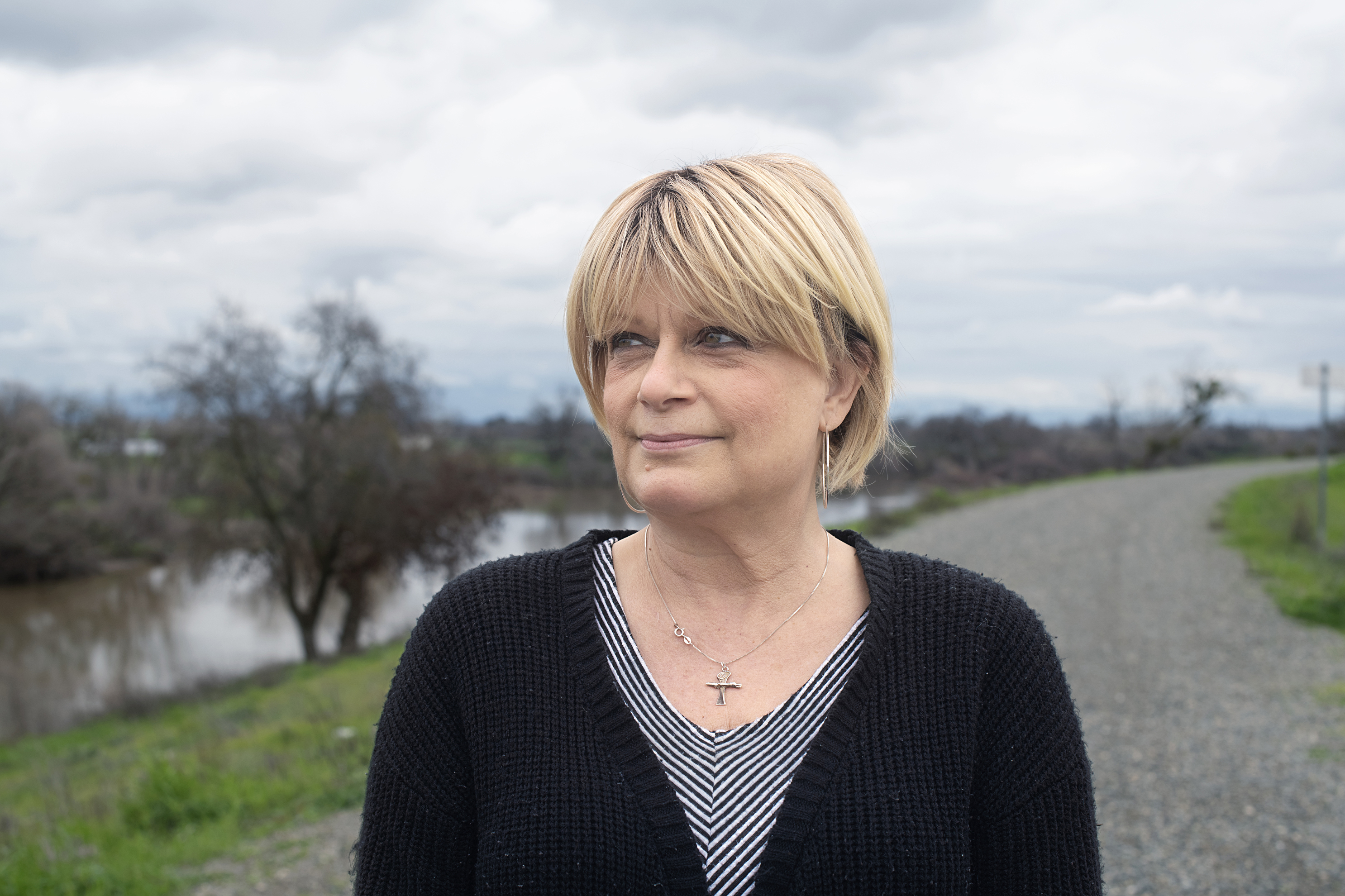 A medium closeup of a woman with short blonde hair wearing a striped tshirt, black cardigan and a silver cross around her neck