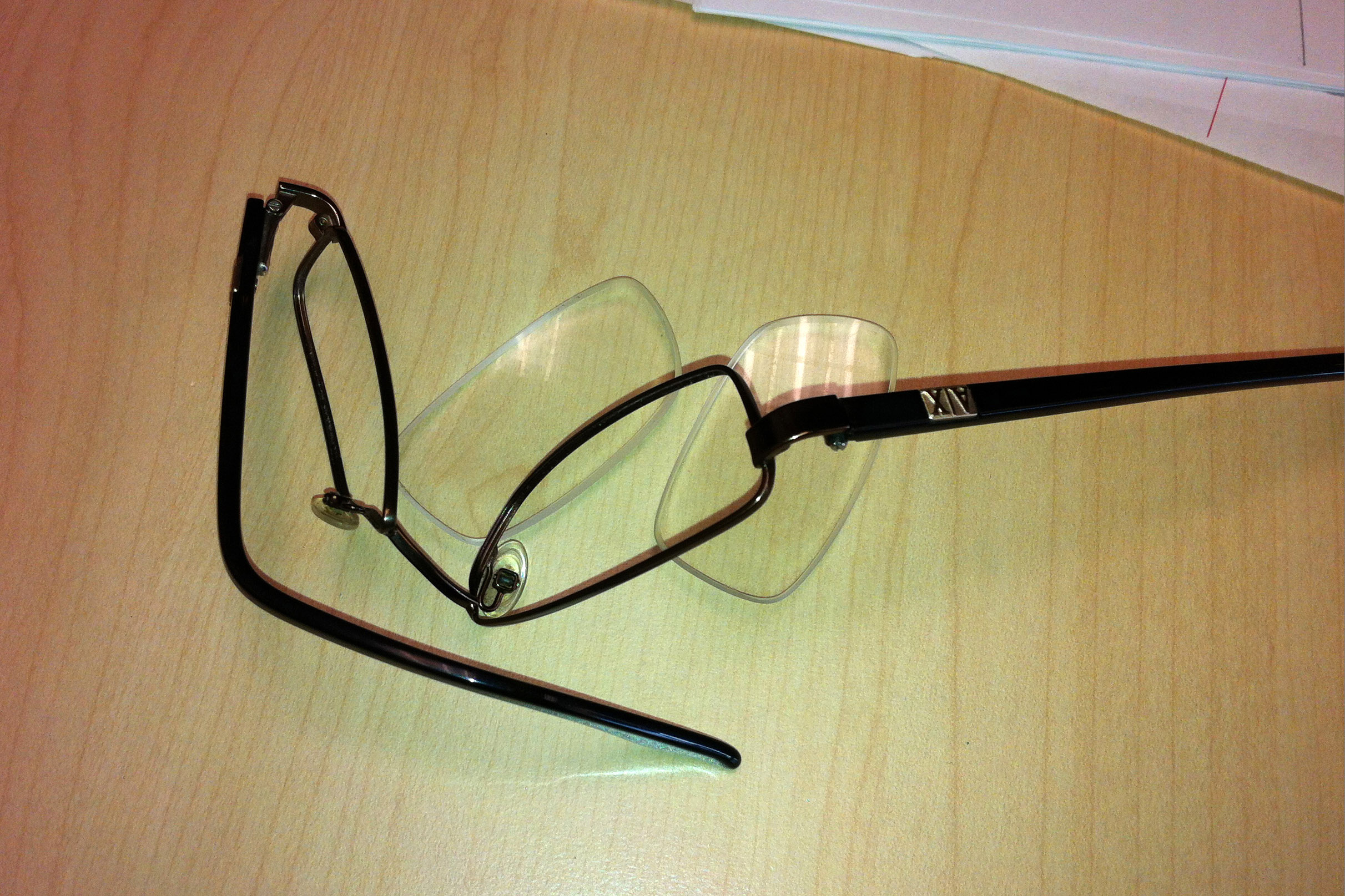 A pair of crumpled, broken glasses are shown on a table. The lenses have been popped out and are next to its warped frame.