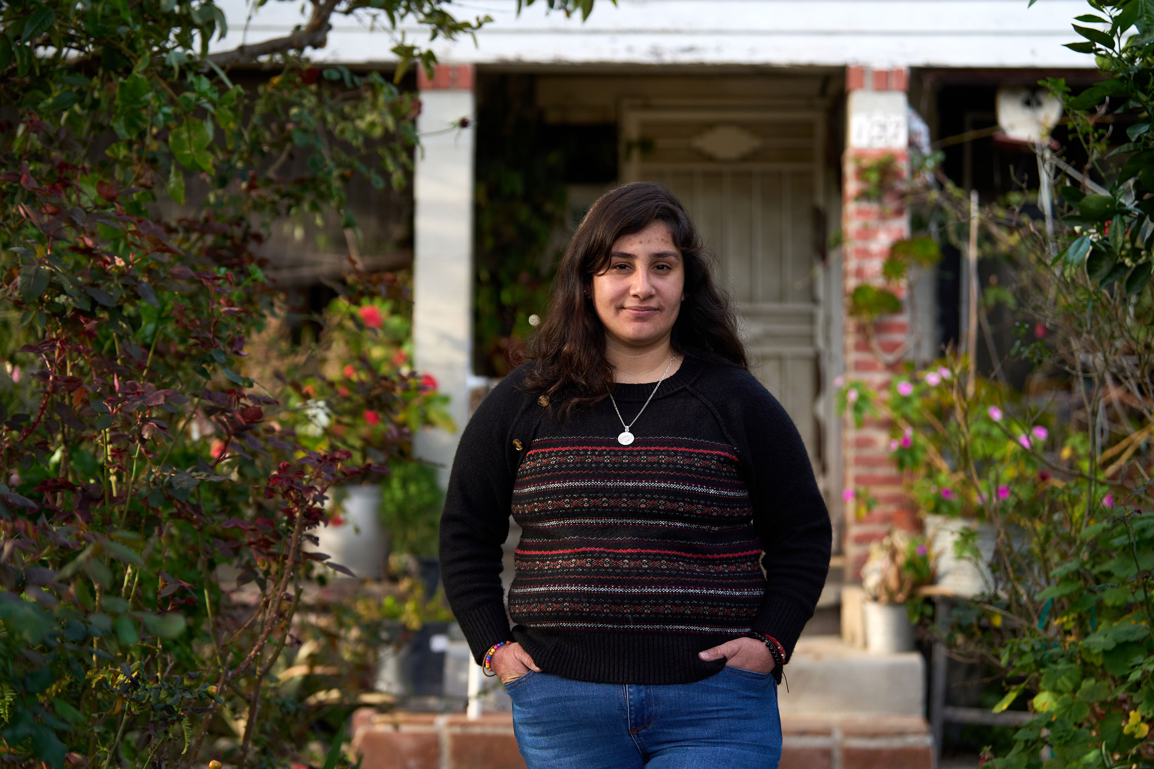 María Rivas Cruz stands in the front walkway of her home. She is centered in the image and looks towards the camera. There are lush, green plants around her on both sides.