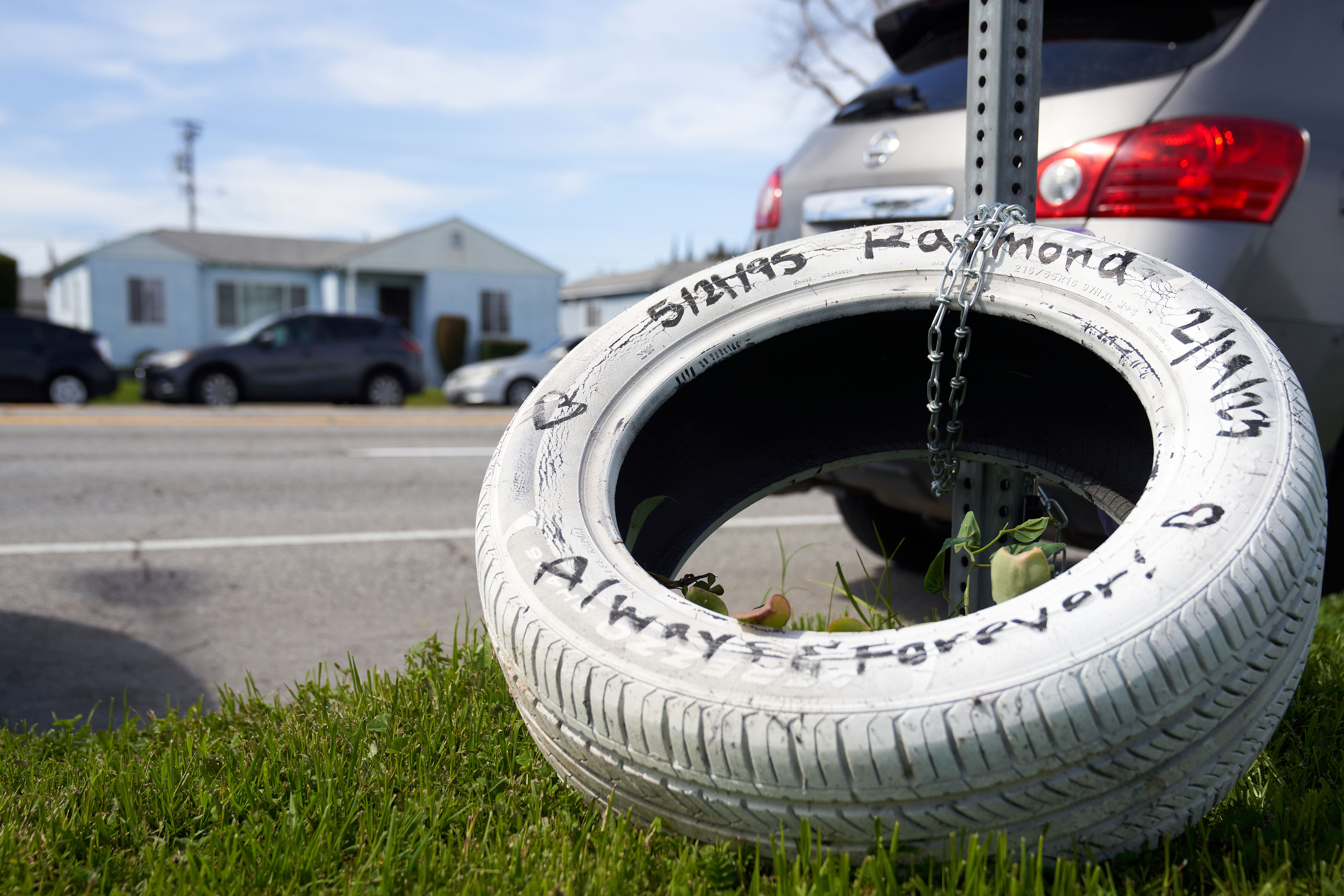 A tire, that has been painted white, is chained to a metal post, as a memorial for Raymond Olivares. The tire has hearts painted on it, and text that says, "5/21/95 / Raymond / 2/19/23 / Always & Forever!"