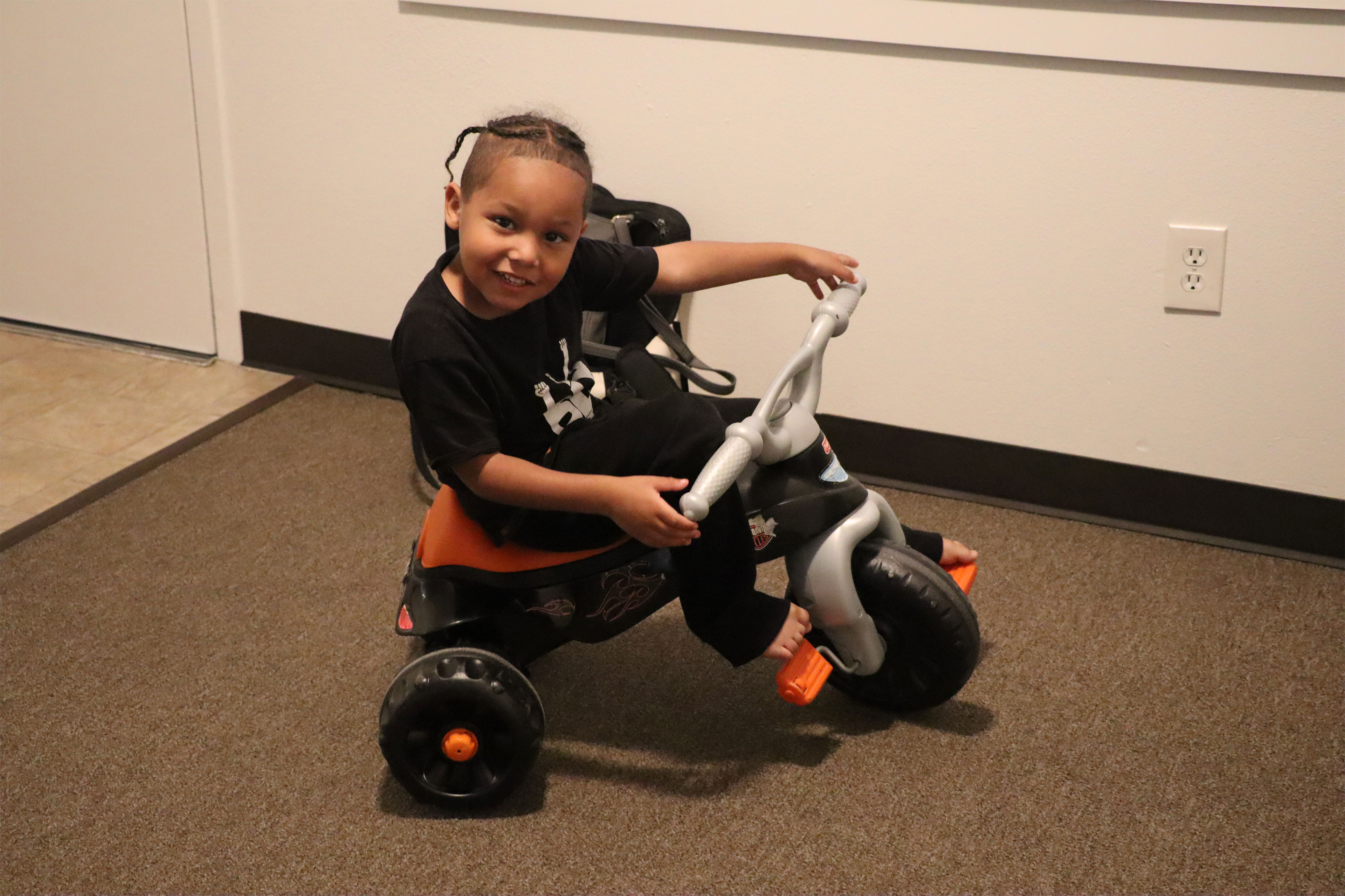 A young boy rides a tricycle inside. He turns and tilts his head toward the camera and smiles.