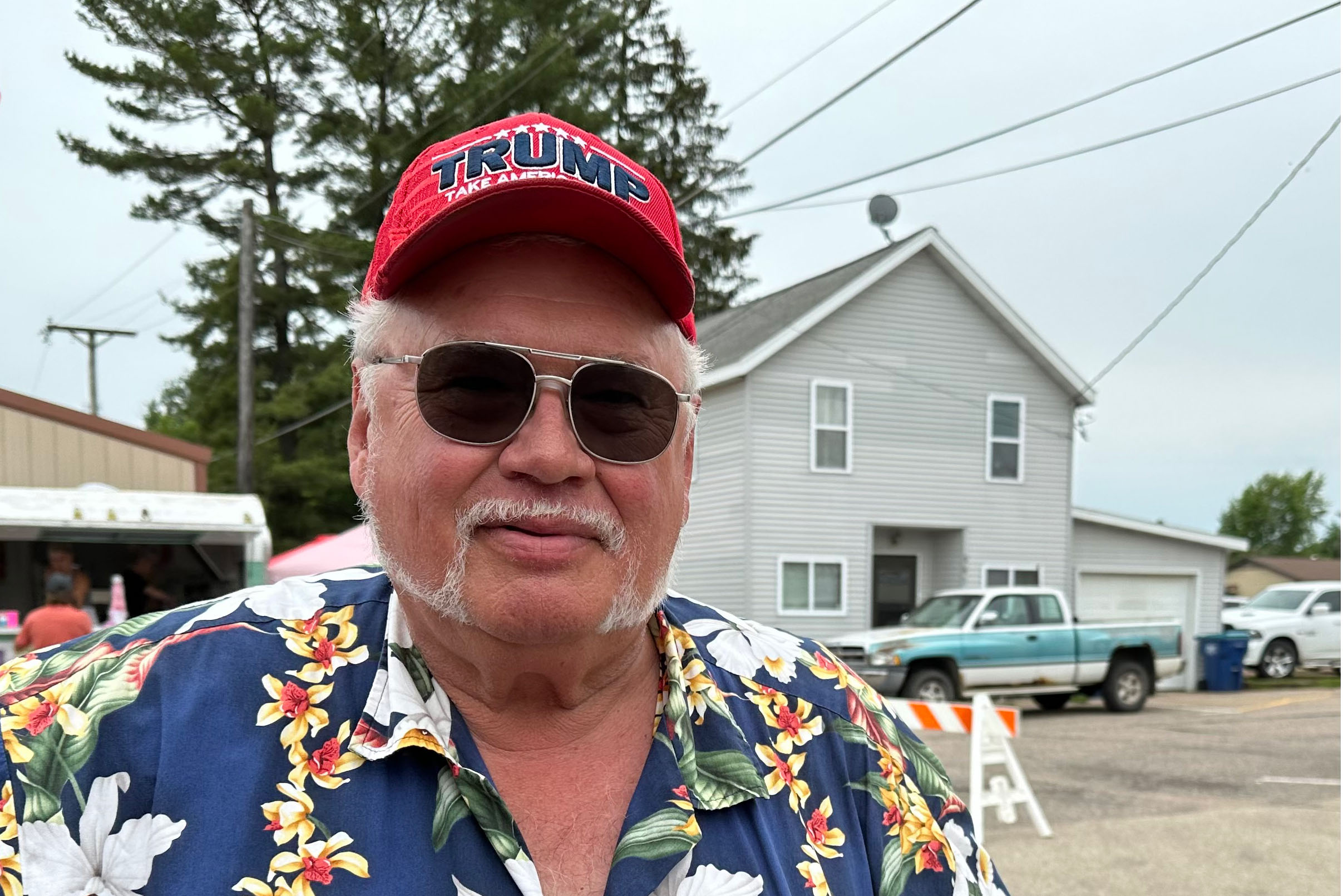 A portrait of Greg Laabs, who is an older man wearing a goatee mustache, aviator sunglasses, and a red hat that says, "TRUMP."