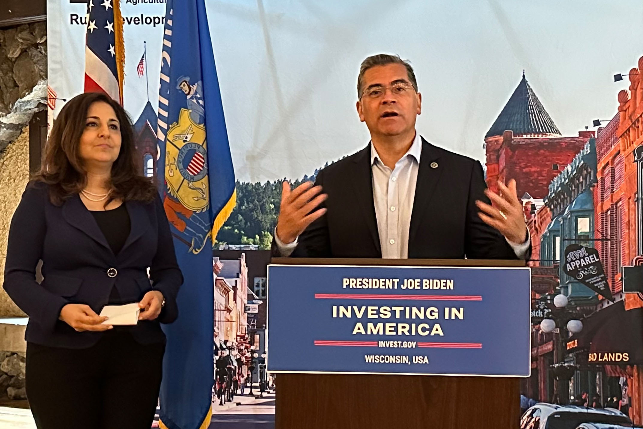 Xavier Becerra stands at a podium that says, "President Joe Biden / Investing in America." Neera Tanden stands beside him.