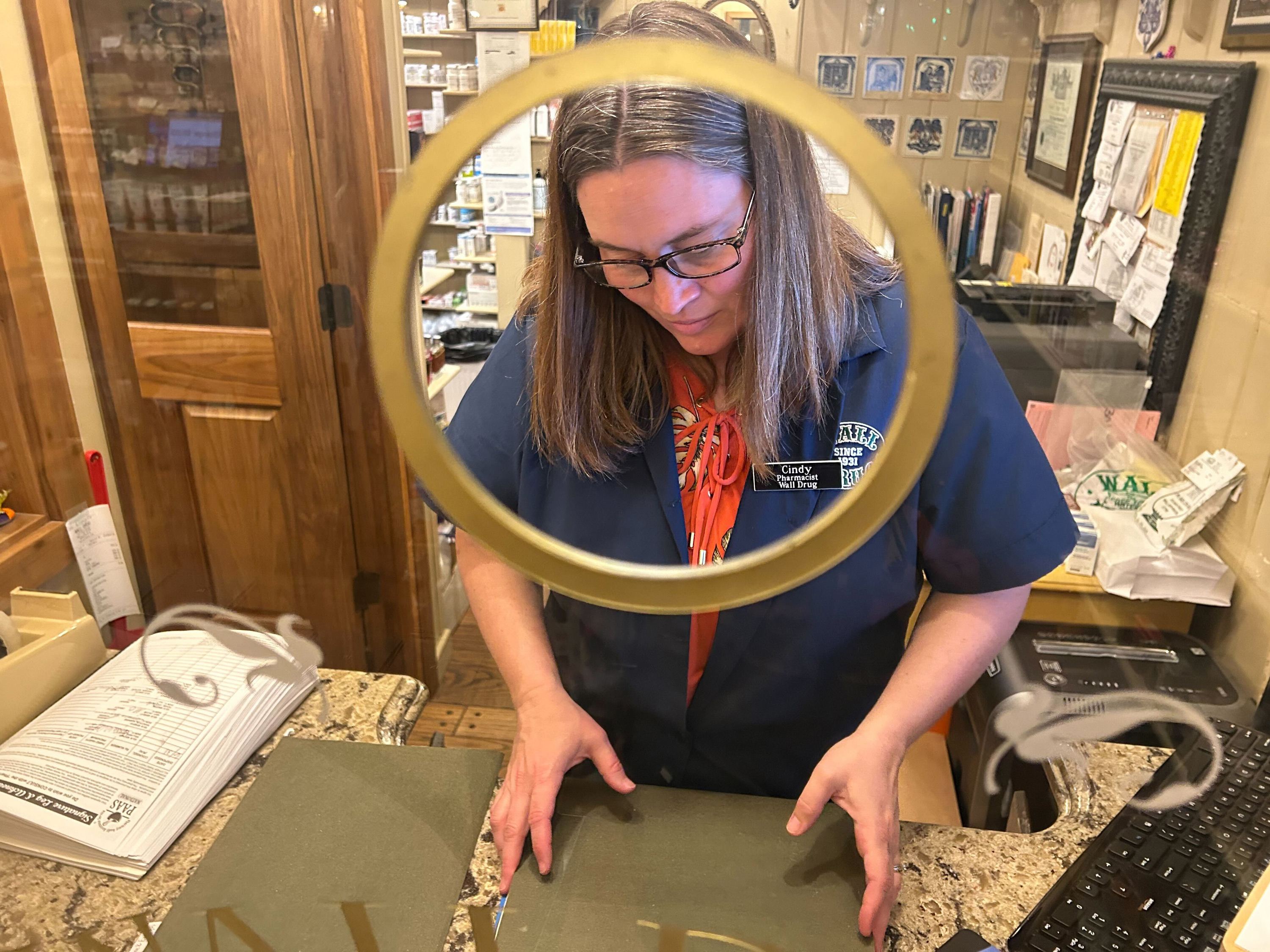 A photo of a pharmacist seem behind a pane of glass with a round hole. The hole frames her head and shoulders.