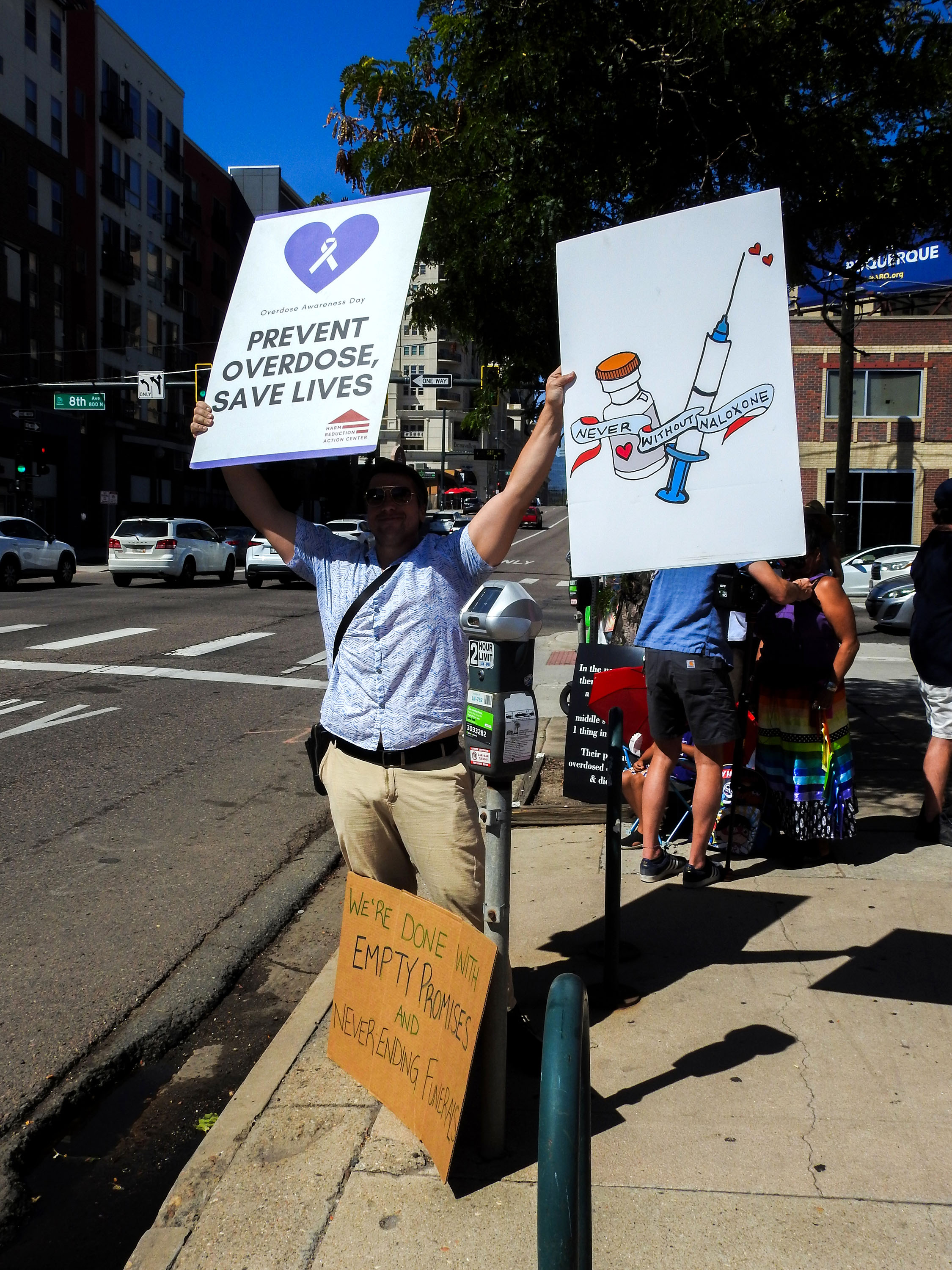 A man is standing on the side of a main road holding up two signs. The sign on the left says: 