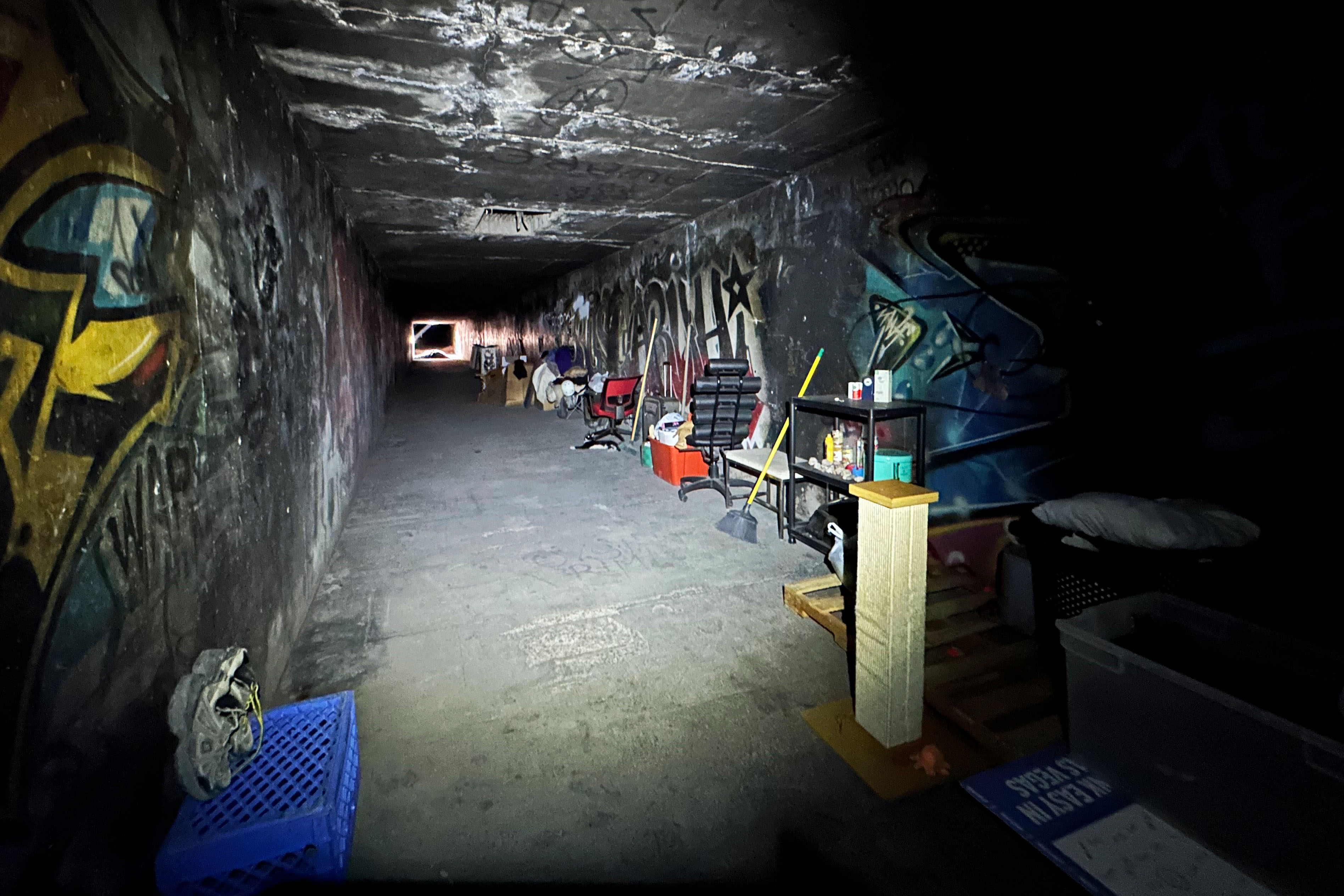 An underground tunnel made of cement. There are personal belongings and other objects near the walls. 