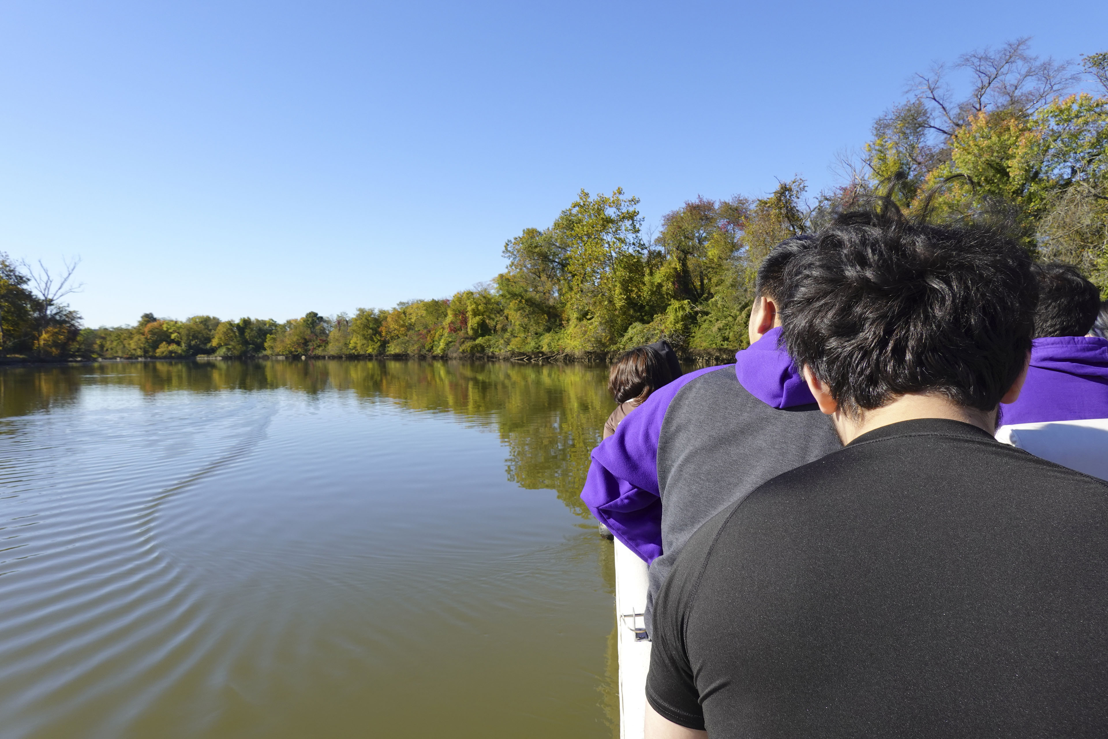 A photo taken from behind a small group of students on a small boat. A river with deep green water stretches out before them. The riverside is lined by green trees. It is a sunny day.