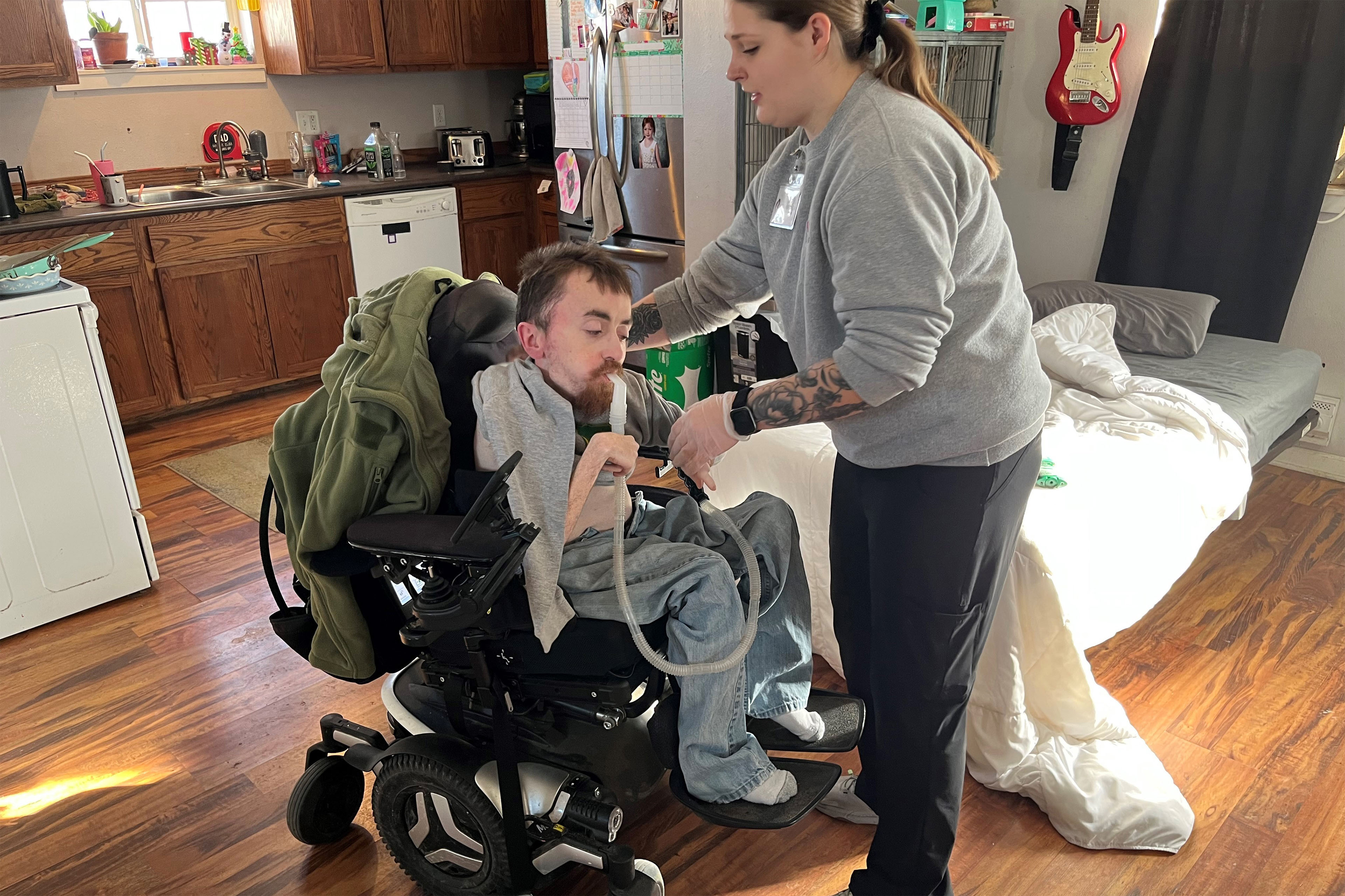 A photo of a man in a wheelchair receiving help putting on a shirt.