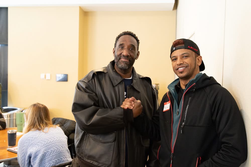 A photo of two men shaking hands and smiling for the camera.