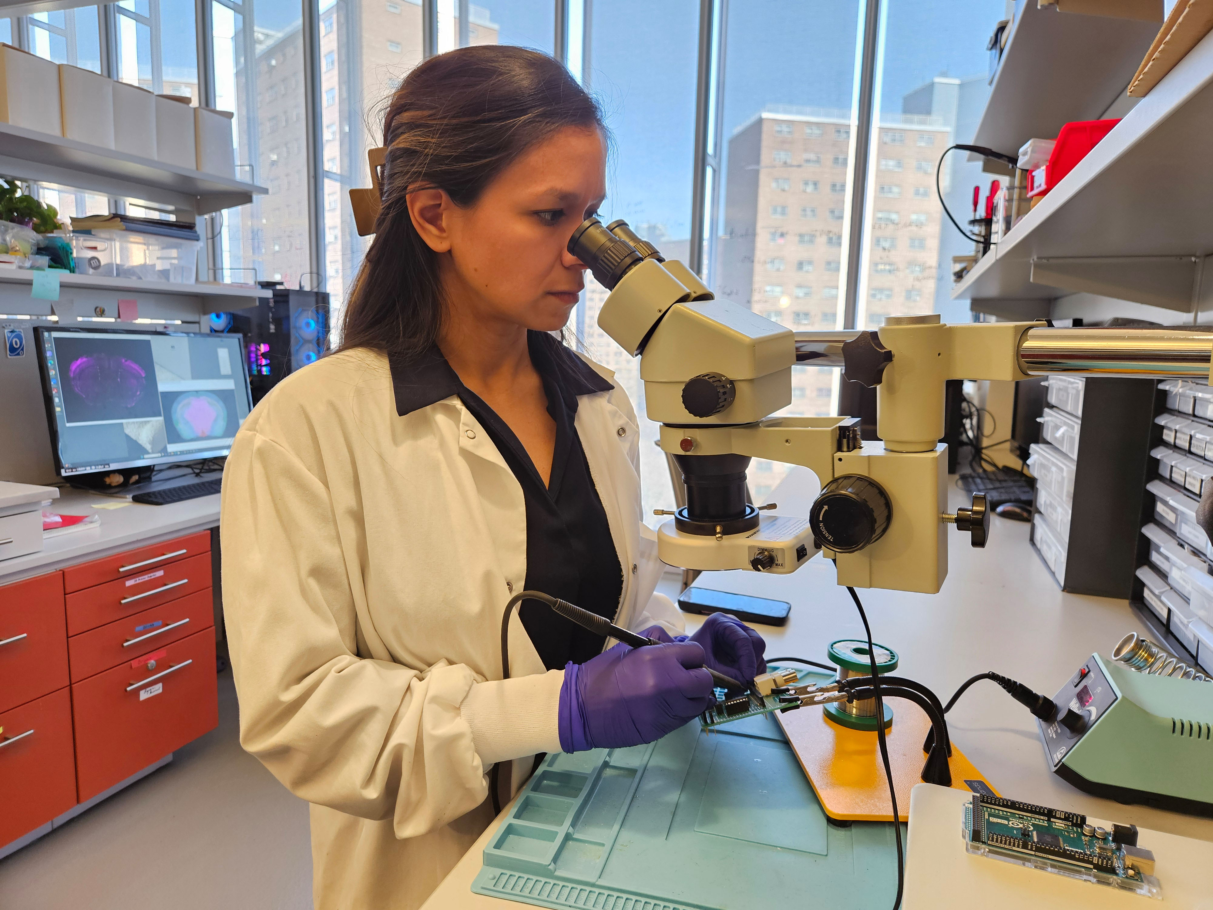 A photo of a woman in a lab coat peering through a microscope while soldering.