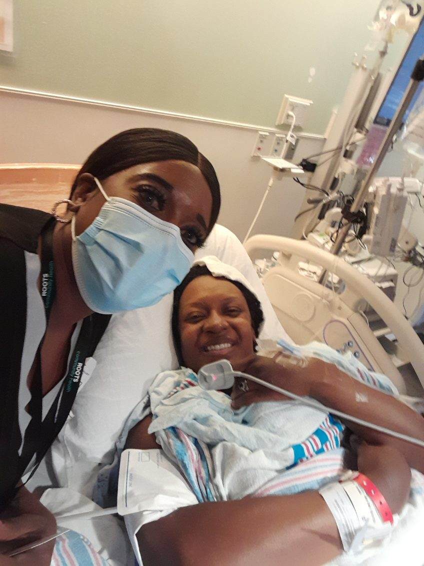 A photo of a doula wearing a mask posing for a picture with Eboni Tomasek lying in a hospital bed, smiling.