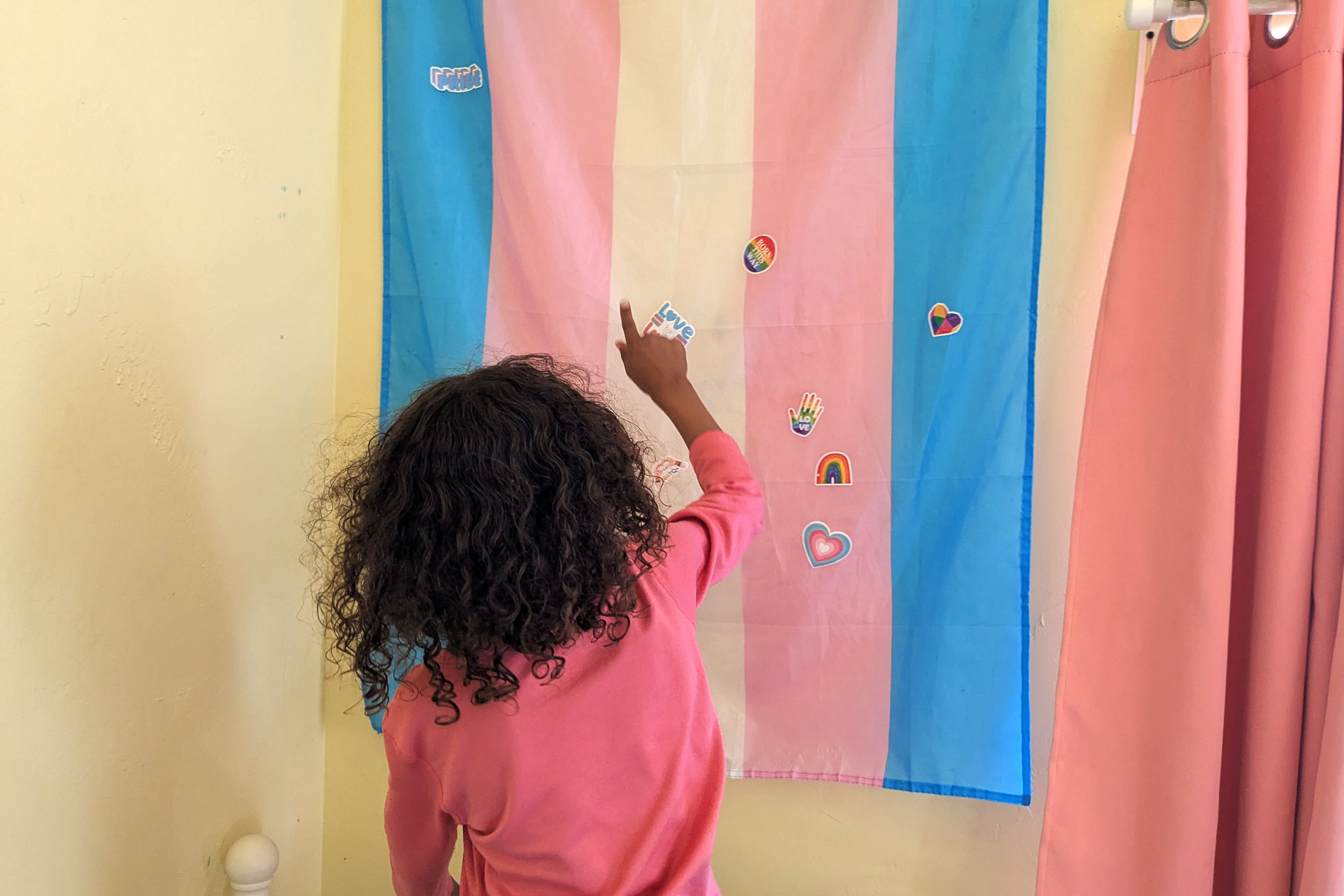 A kindergarten-aged child with curly brown hair stands in front of a transgender flag that has rainbow and heart-shaped stickers on it. The child points up at the flag.