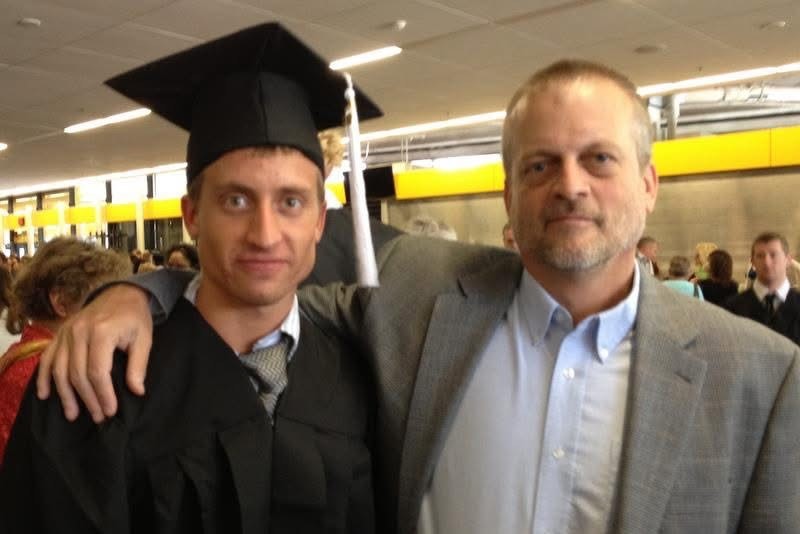 A photo of a man in a graduation cap and gown posing for a photo with his father, who has his arm wrapped around his son.