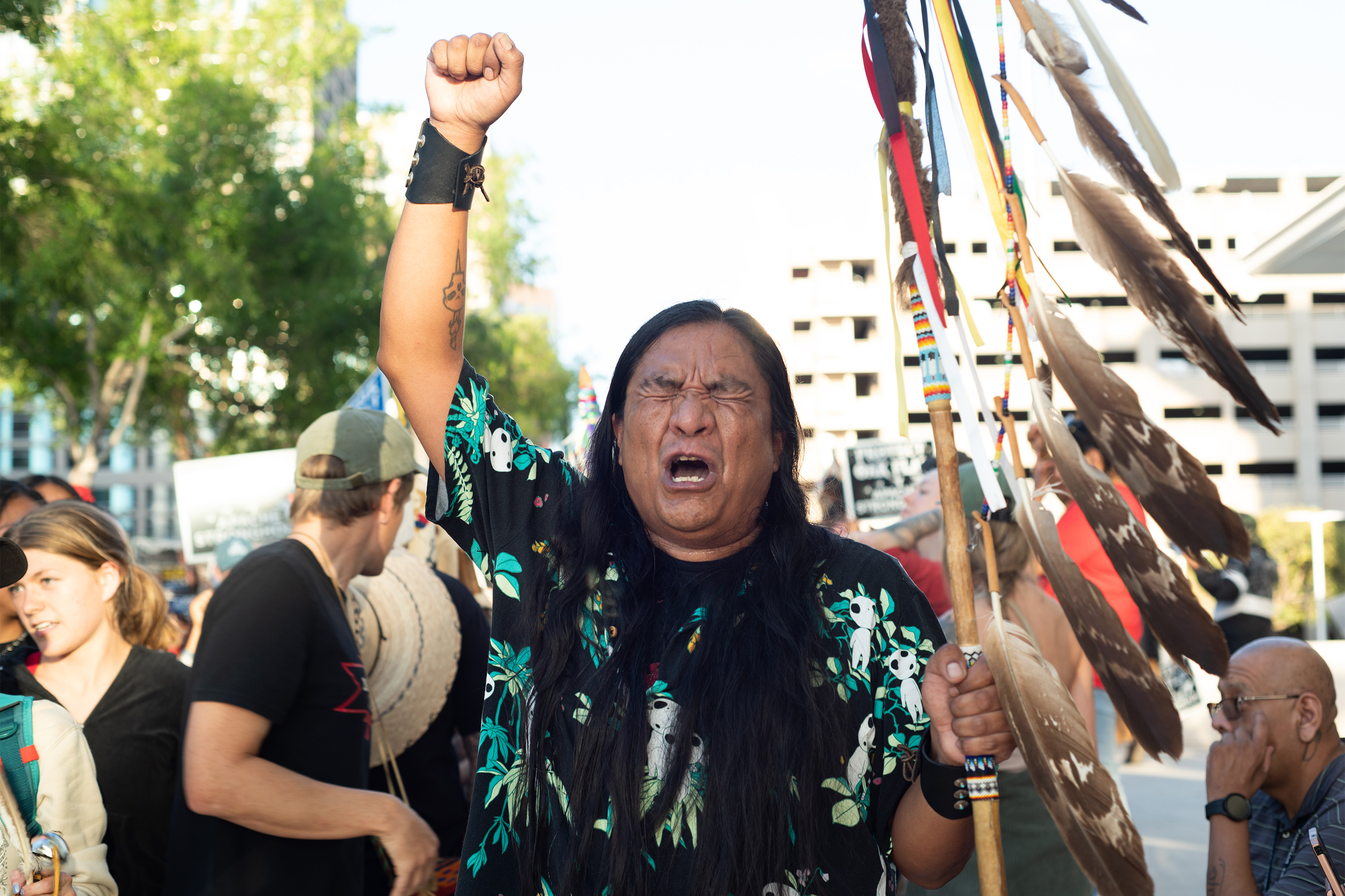 A photo of an indigenous man raising a fist and a staff. He chants with his eyes closed.
