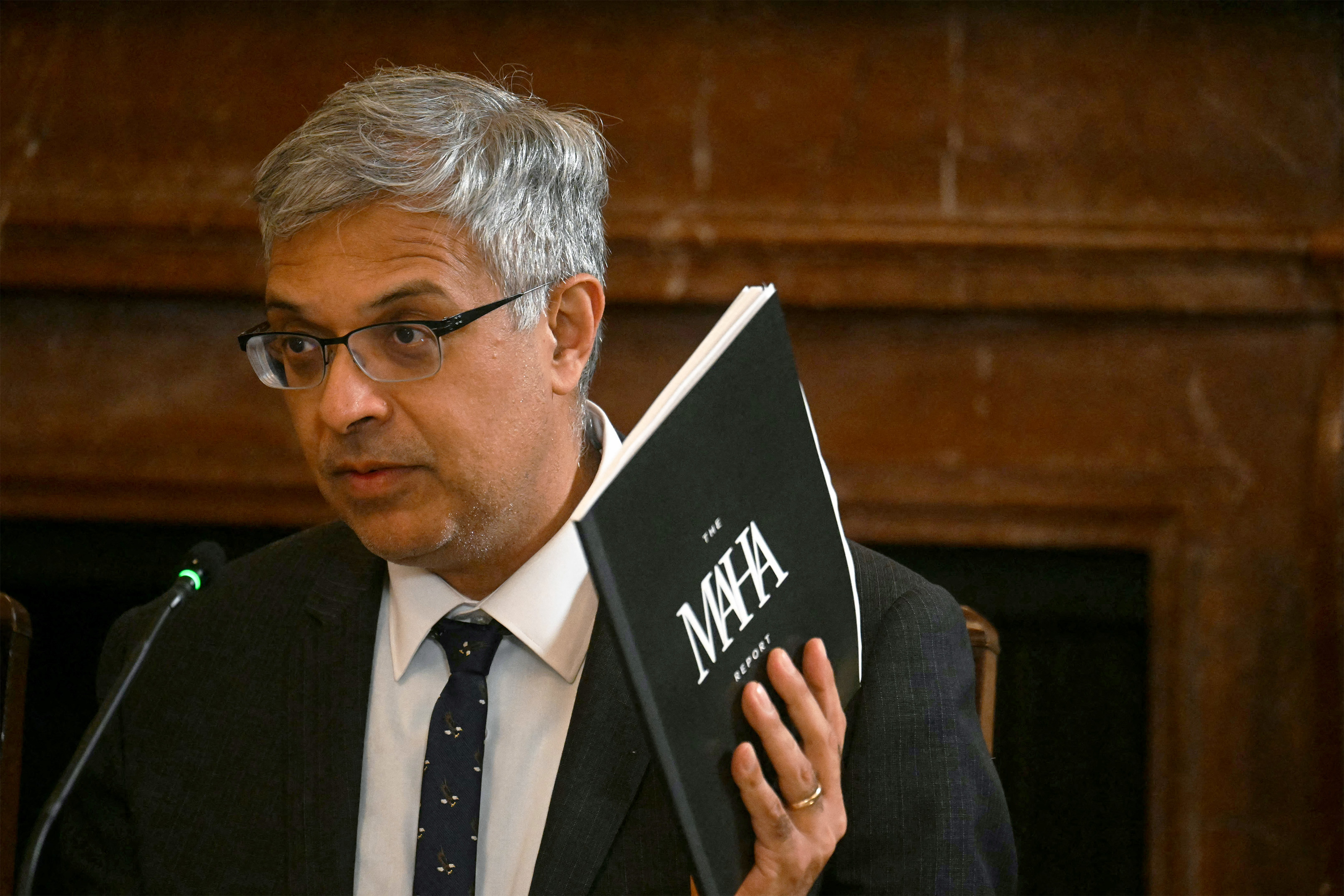 A photo of Jay Bhattacharya seated at a table, holding a printed booklet with a black cover. The cover reads, "The MAHA Report."