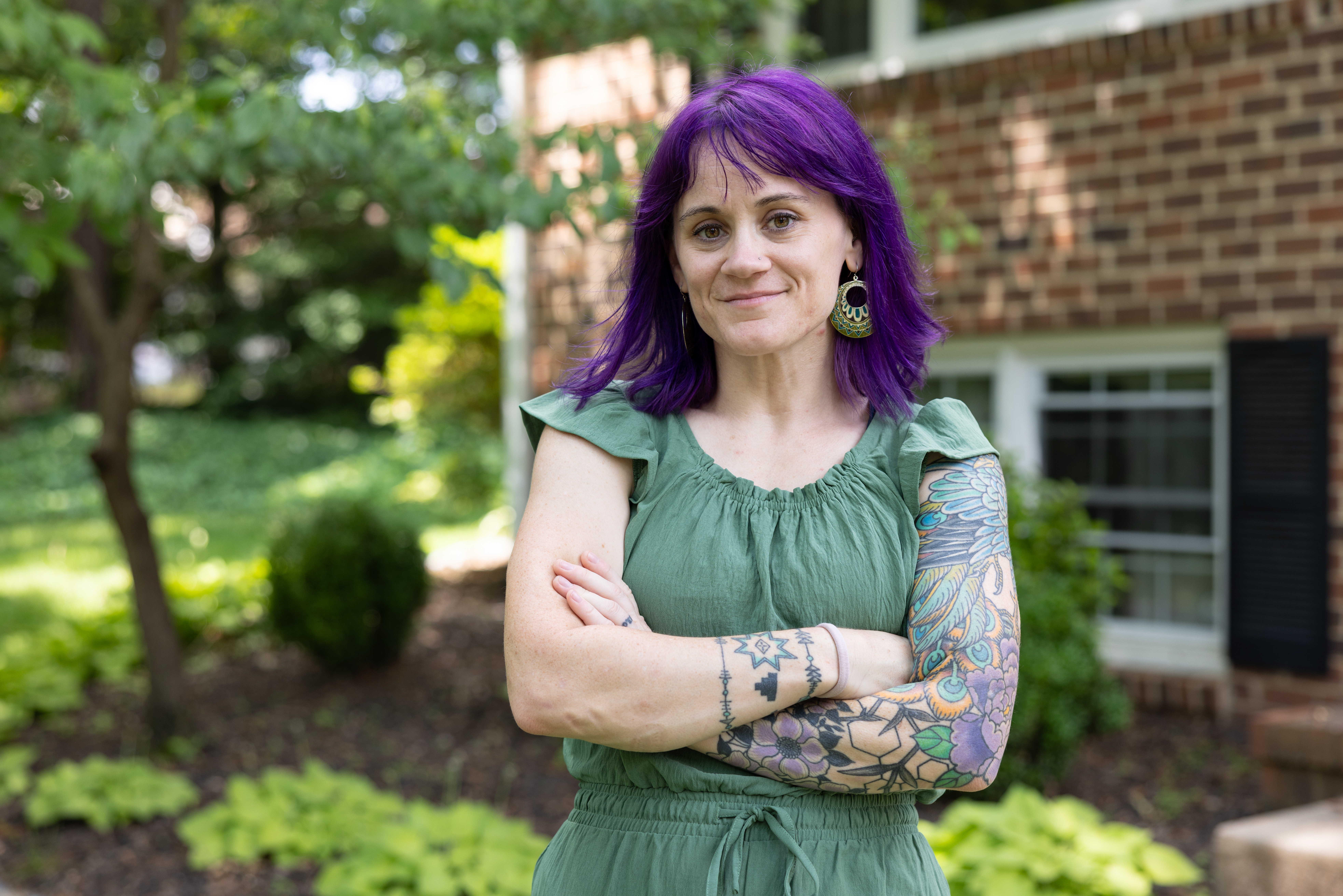A woman with bright purple hair is wearing a green dress and standing outside with her arms crossed. 