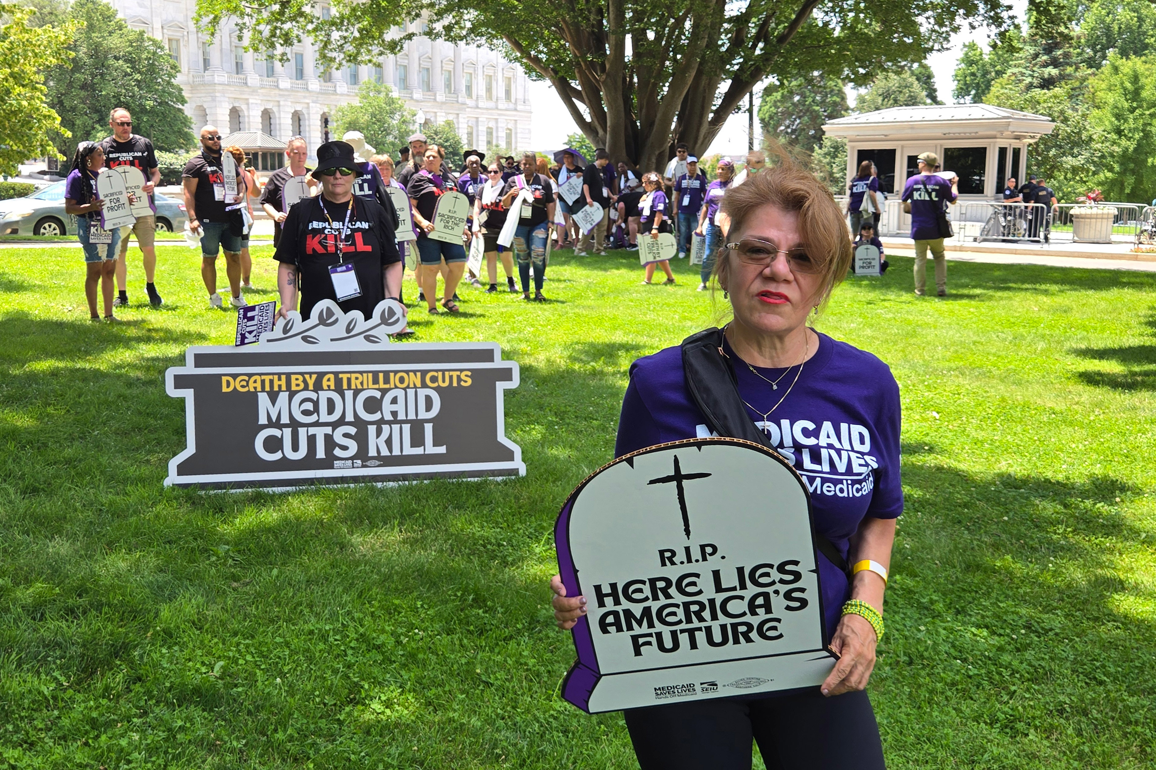 A photo of a woman standing for a portrait outside. She holds a protest sign shaped like a tombstone reading, 