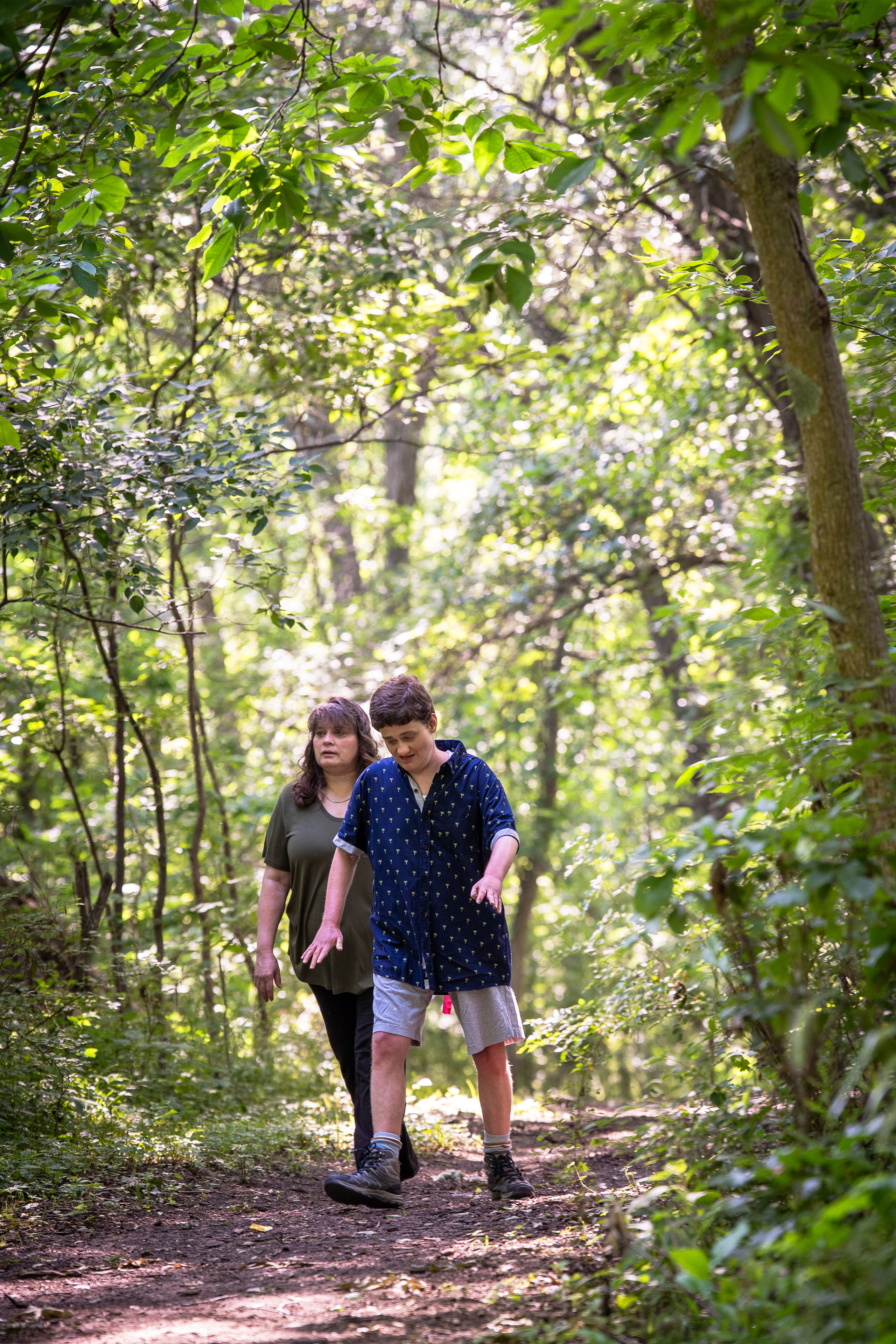 A young man wearing a short-sleeve blue shirt with small palm trees on it and shorts, and a woman wearing an olive green t-shirt and long dark pants, walk along a path surrounded by trees.