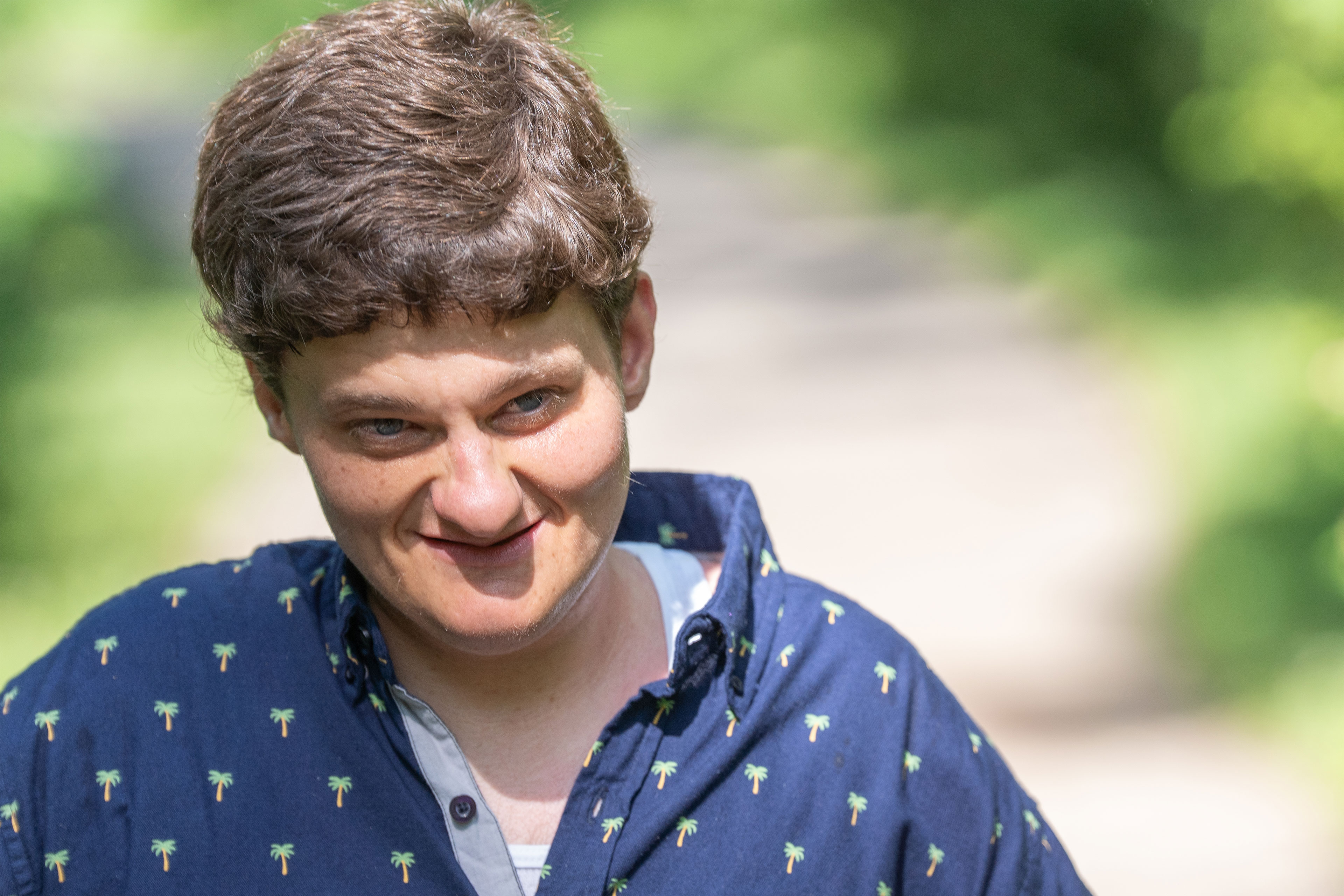 A close-up portrait of a young man with brown hair wearing a blue shirt with palm trees on it stands outside. 