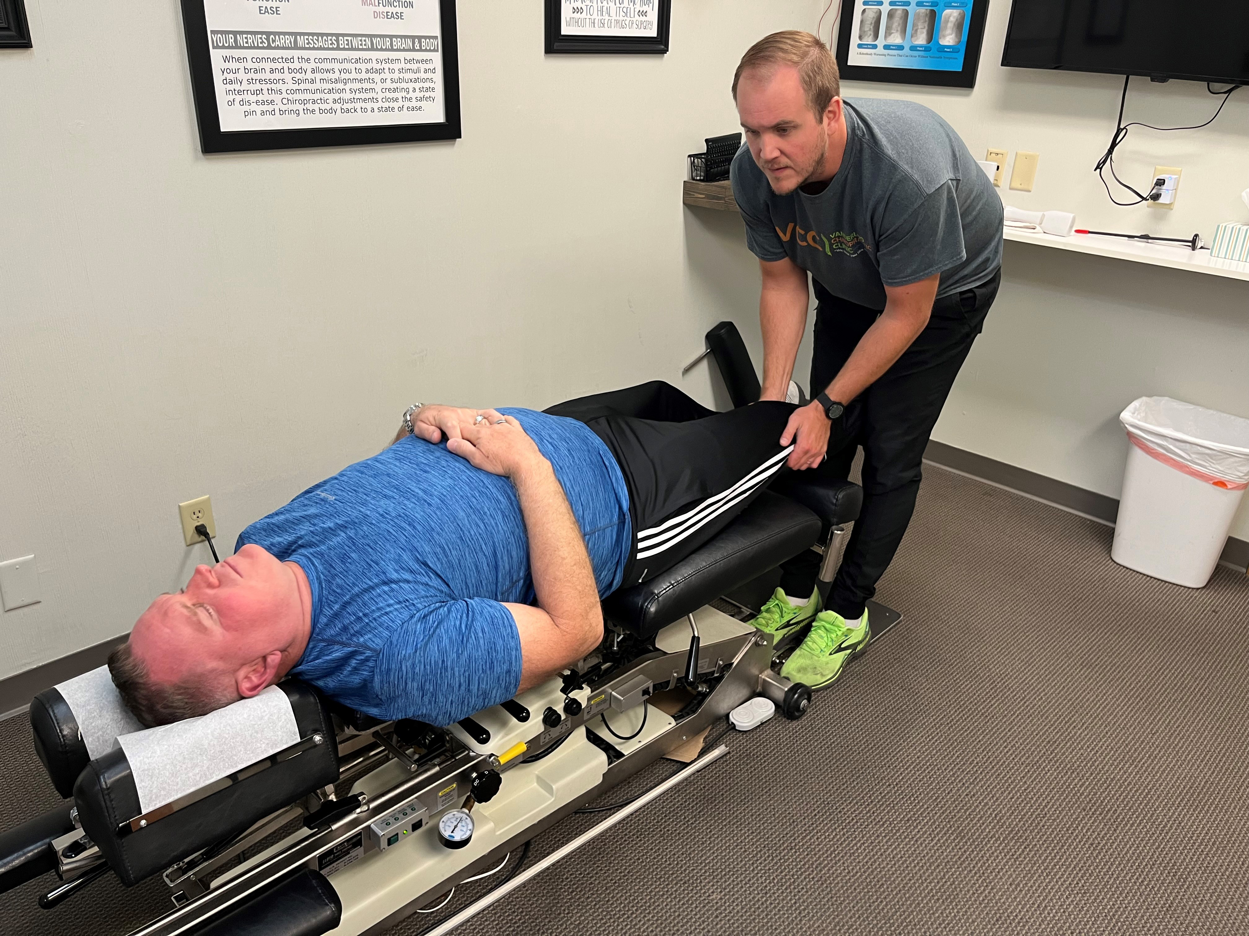 A man in a blue tshirt lays on a chiropractor bed while the chiropractor, in a gray tshirt, leans over his legs
