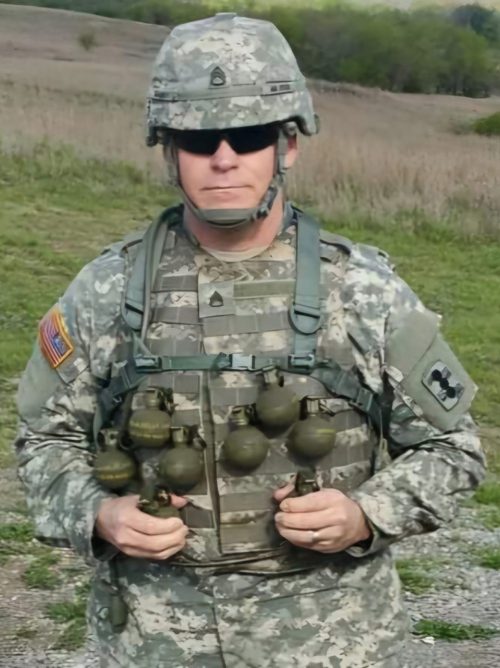 A white man in a camouflage military uniform, including a helmet and sunglasses, stands outside.