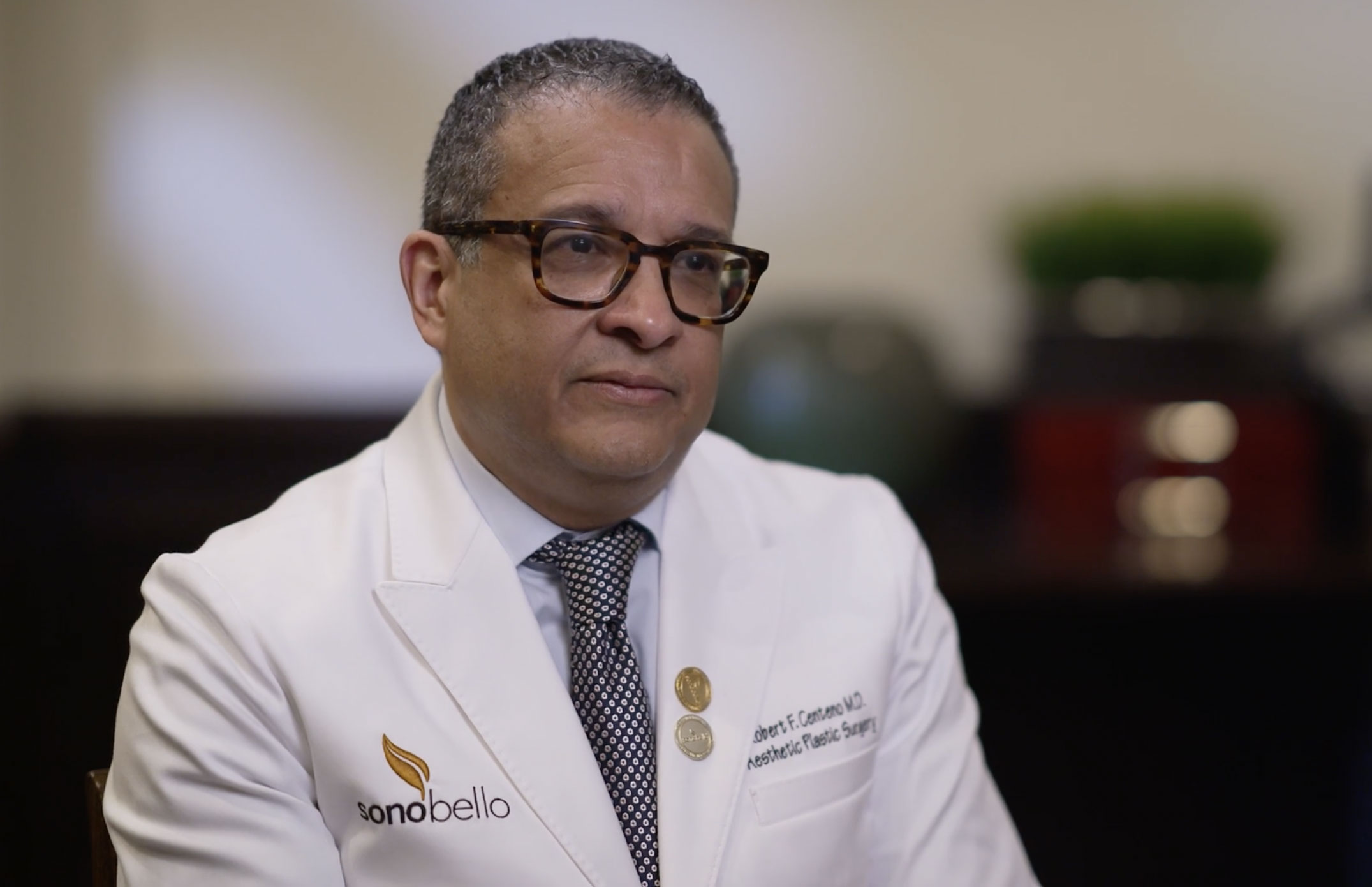 A man wearing a white doctor's coat with the logo for Sono Bello sits in an office.