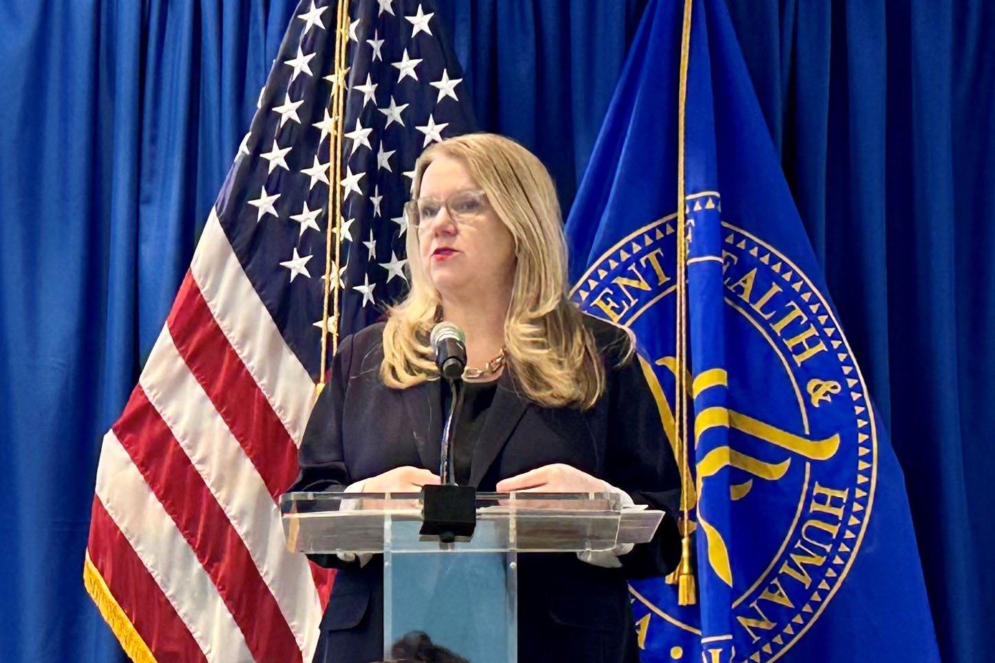 A woman with blonde hair wearing a navy blouse stands at a podium. The American flag and a flag for the Department of Health and Human Services are visible behind her.