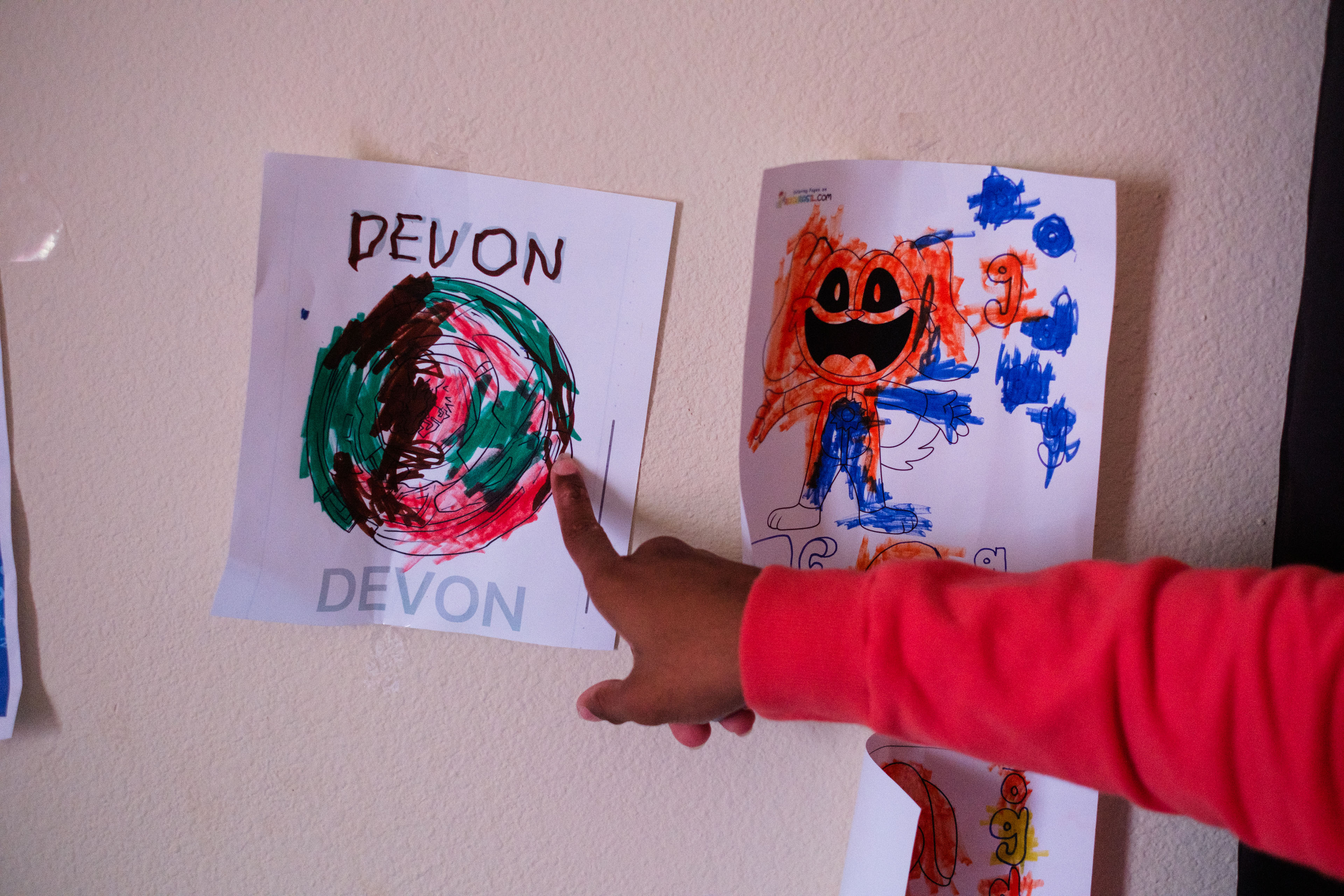 A child's artwork is taped to a wall. The child points to it.