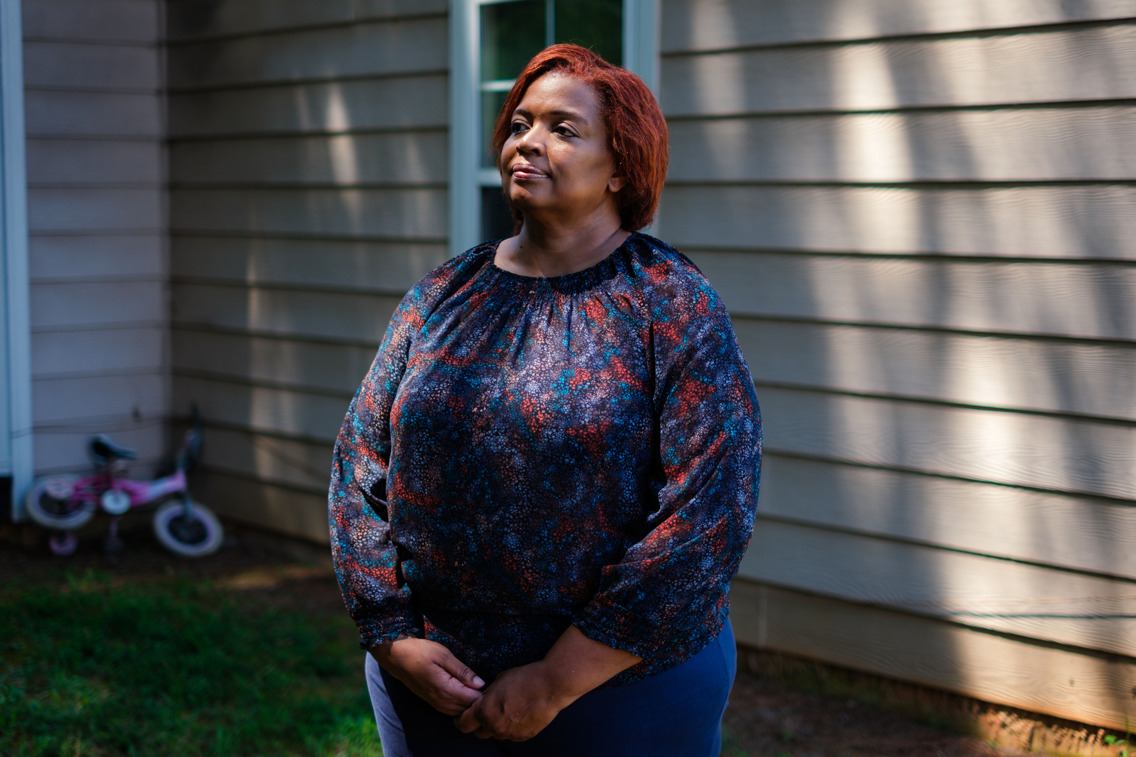 A portrait of a woman standing outside in dappled sunlight. She looks off into the distance.