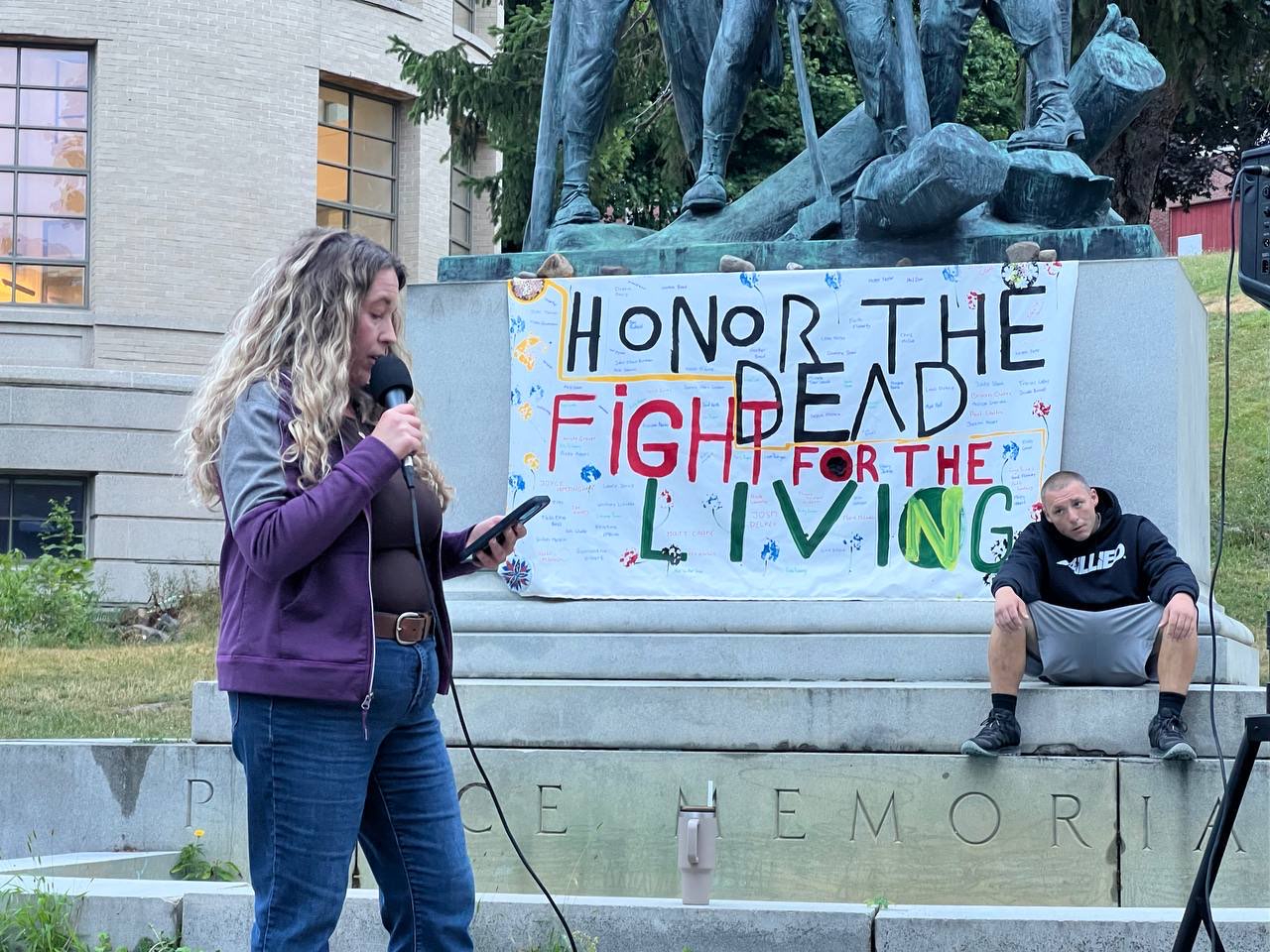 A photo of a woman speaking outside at a rally. A sign behind her reads, "Honor the dead, fight for the living."