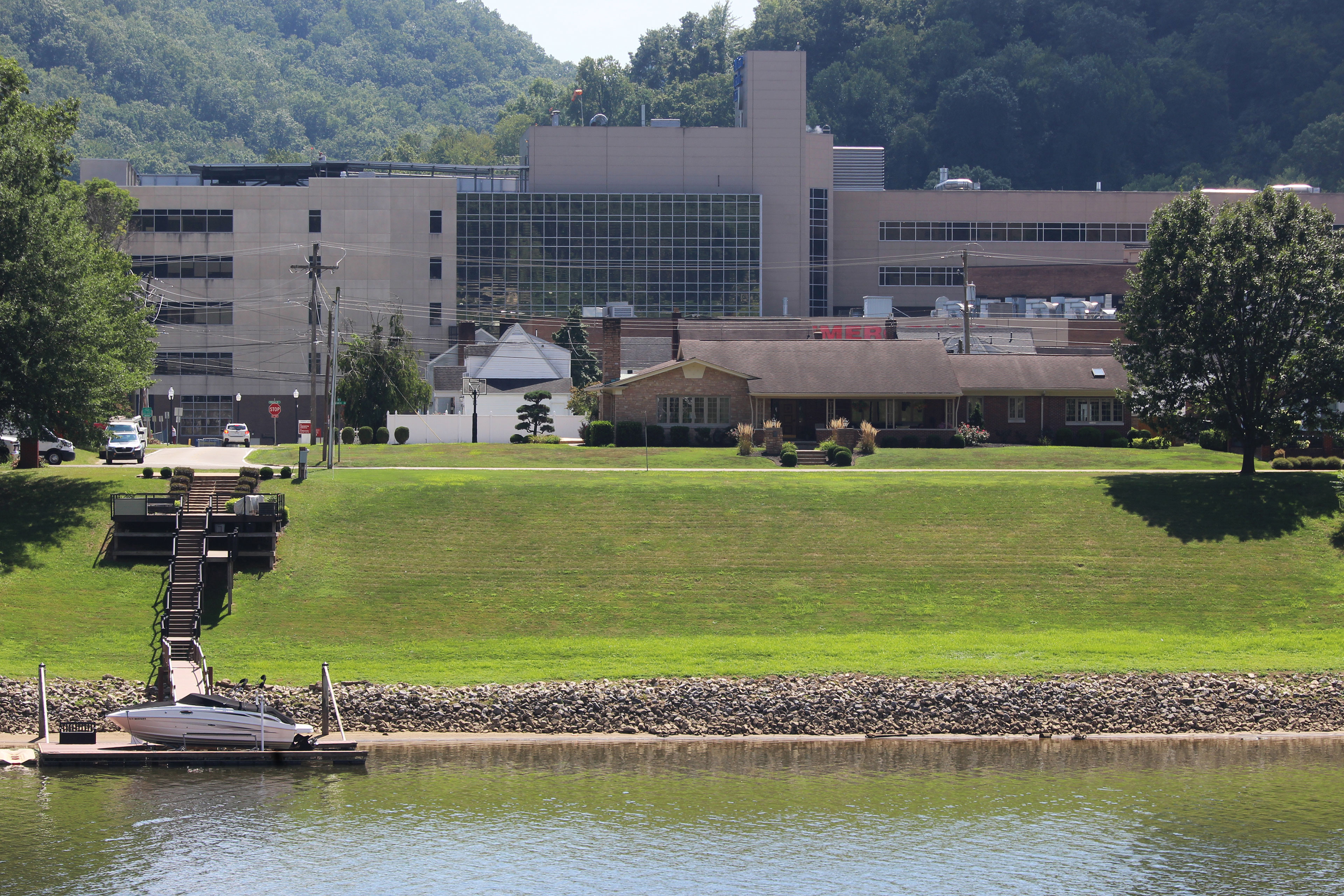 Several buildings can be seen a short distance from a river, beyond a road and some houses. Steps lead down the riverbank to dock and boat.
