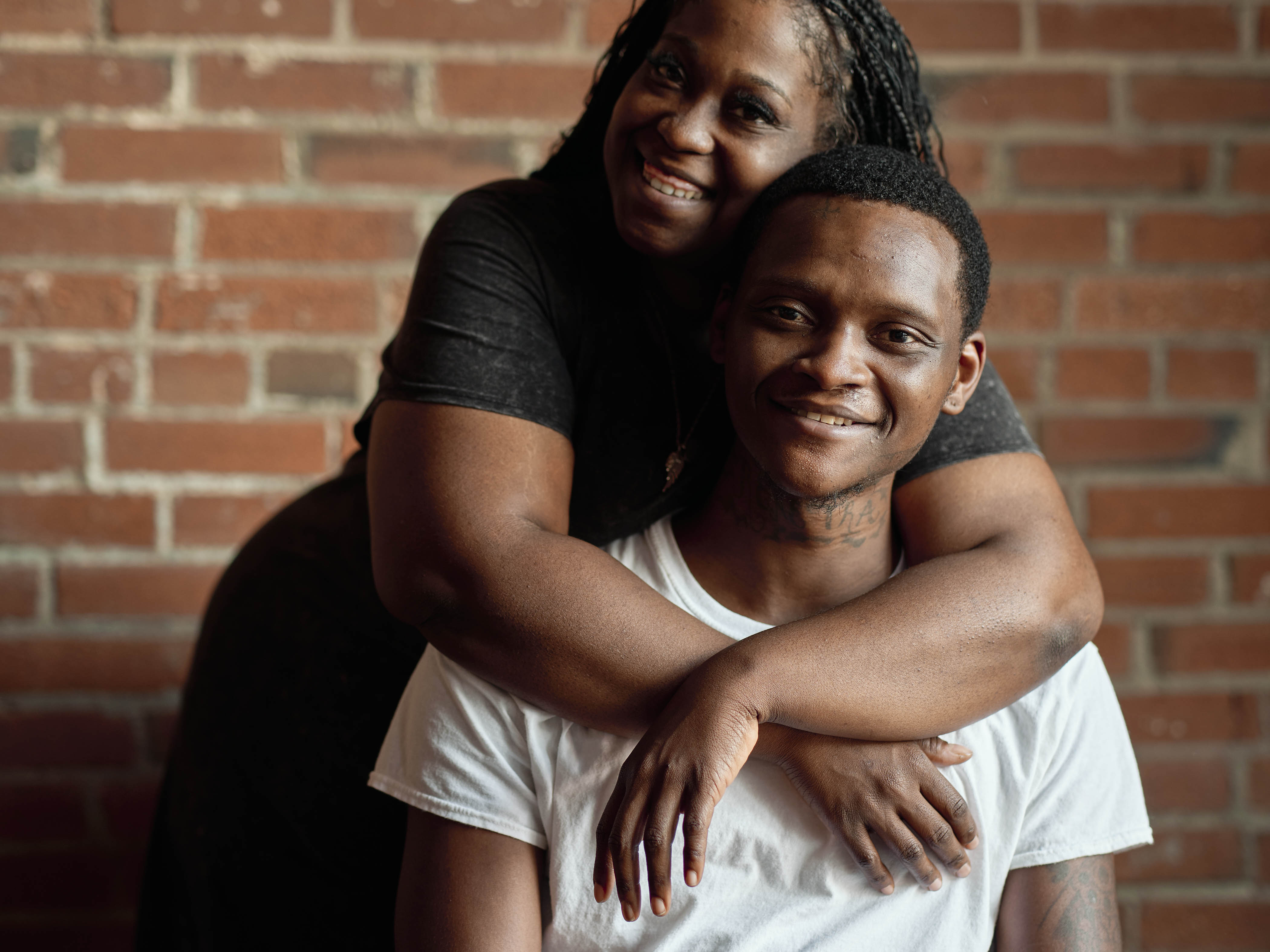 A woman in a black dress drapes her arms around a young man in a white tshirt 