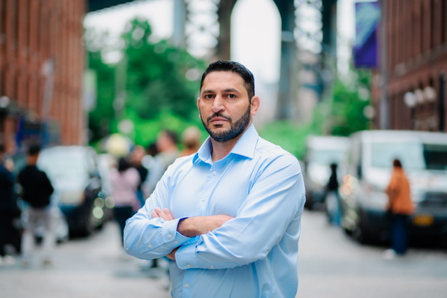 A man with a dark hair and beard in a blue shirt stands with his arms crossed