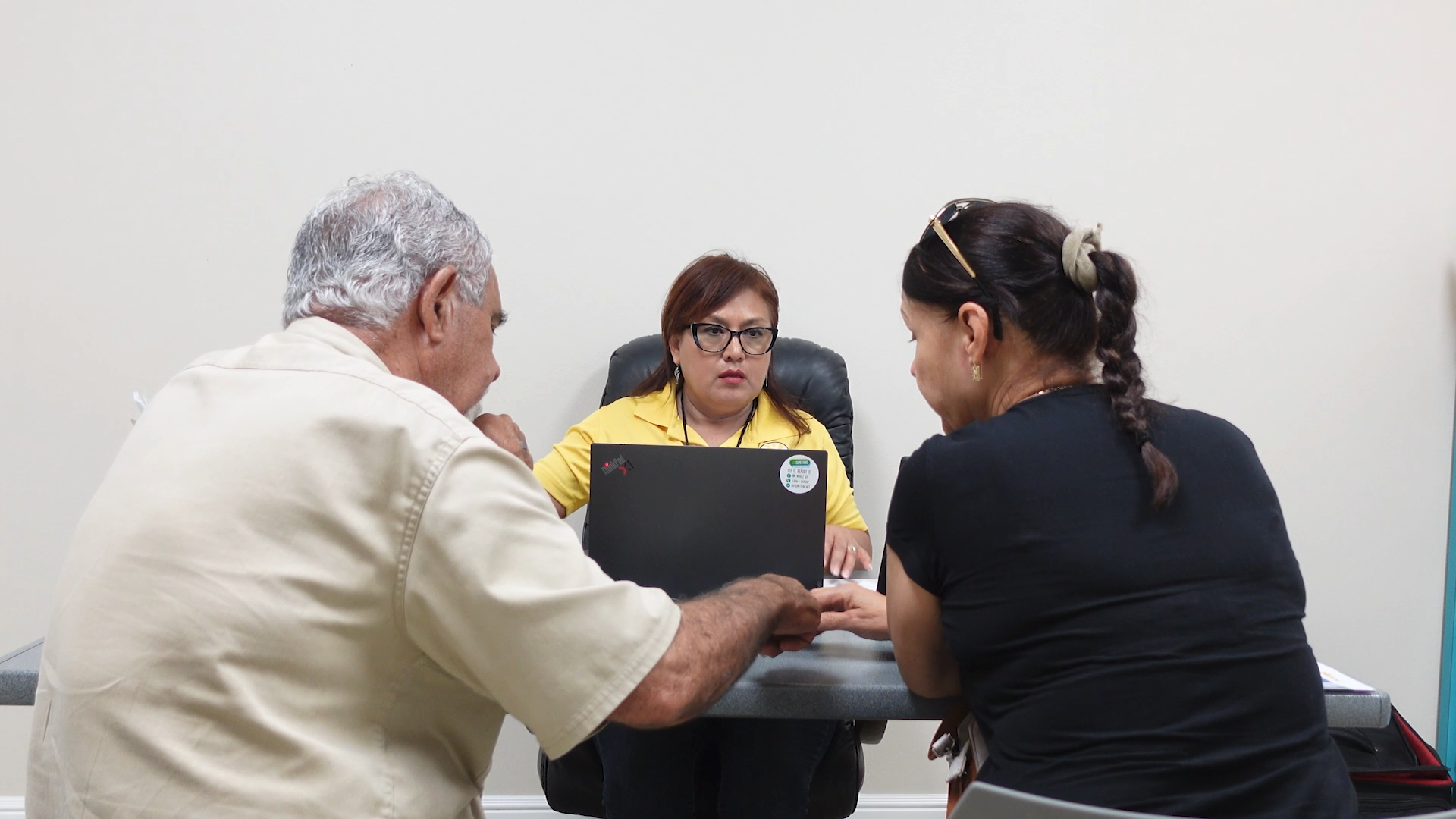 A photo of a promotora sitting behind a laptop in a library meeting room. She's speaking to two clients: an older man and a younger woman.