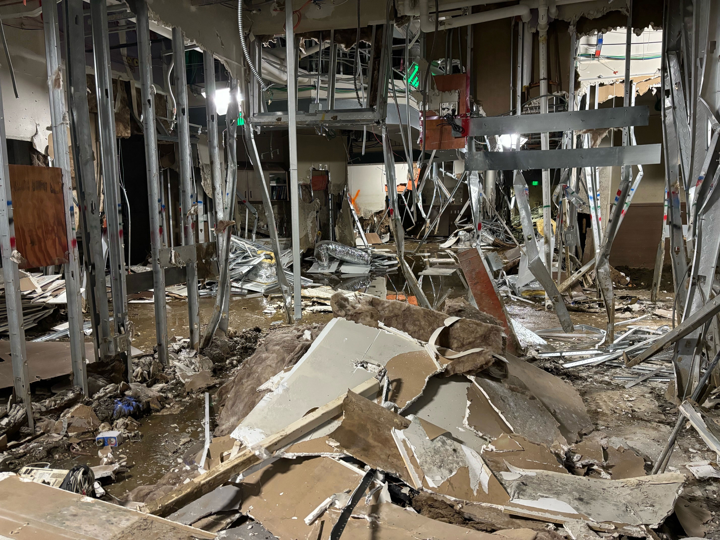 The interior of a destroyed build where metal hangs from the ceiling and drywall, debris, and mud cover the floor.