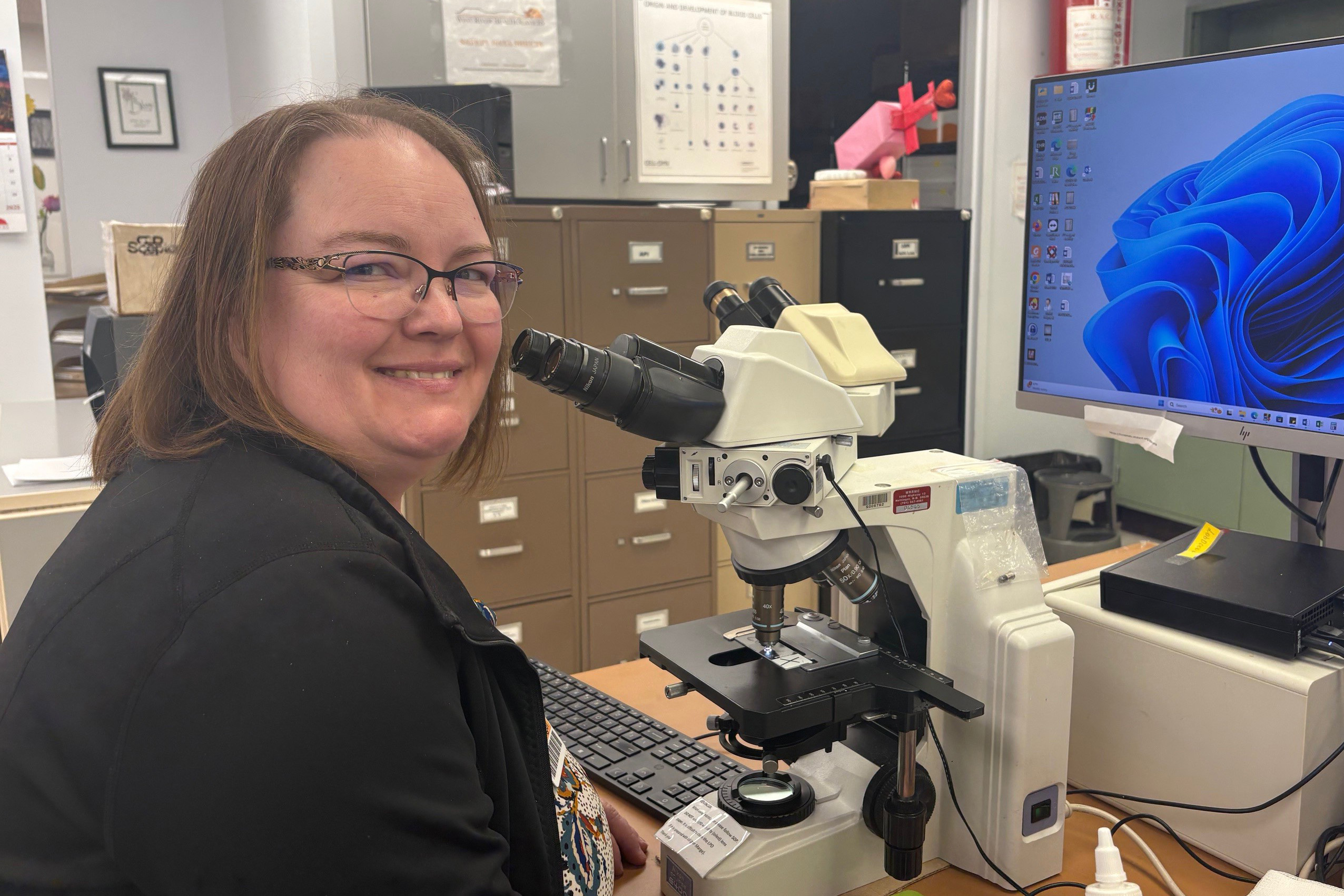 A photo of a white woman sitting in front of a microscope and computer.