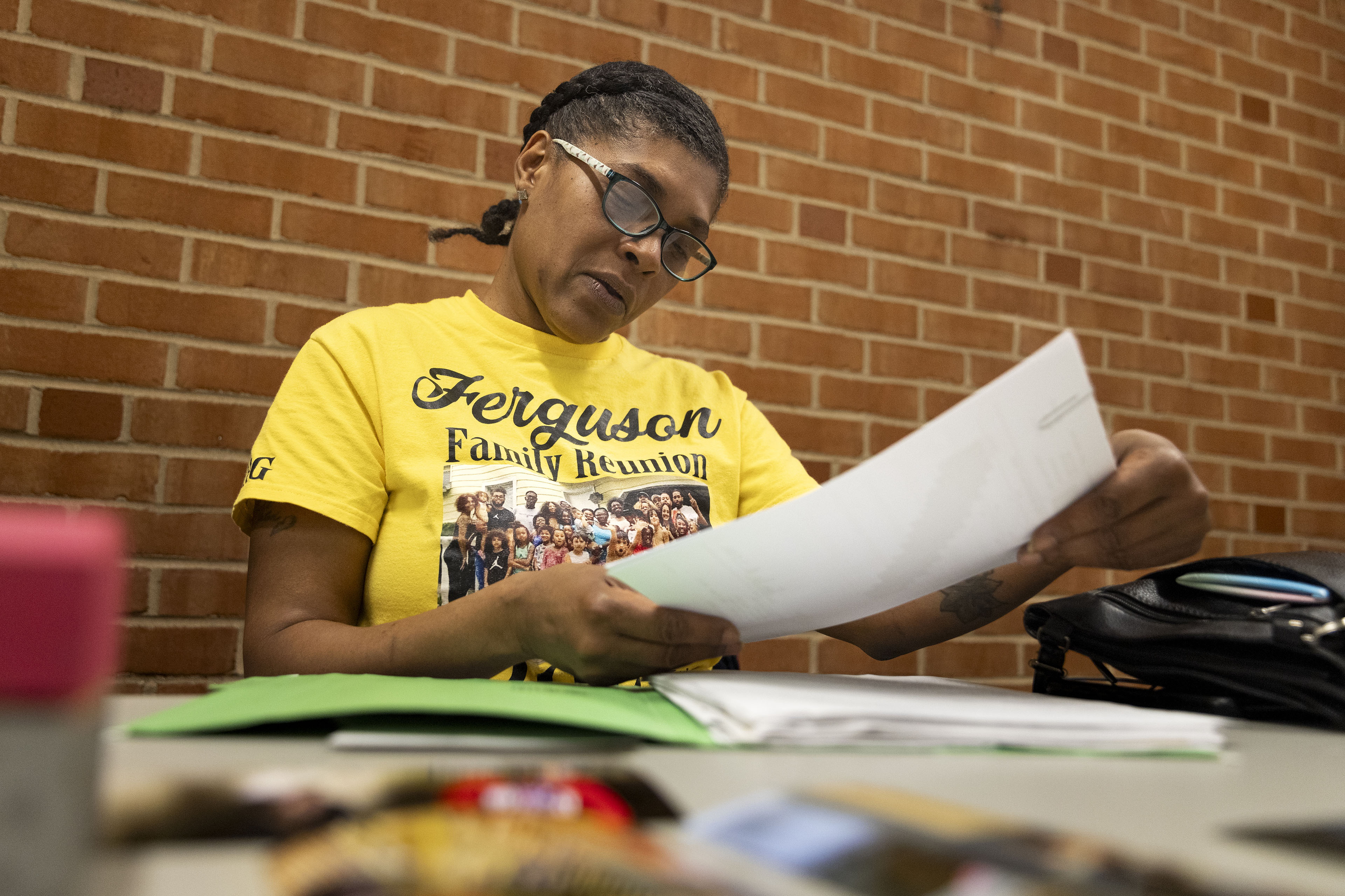 A woman wearing glasses and a shirt that shows her family at a reunion is looking over documents on a table.
