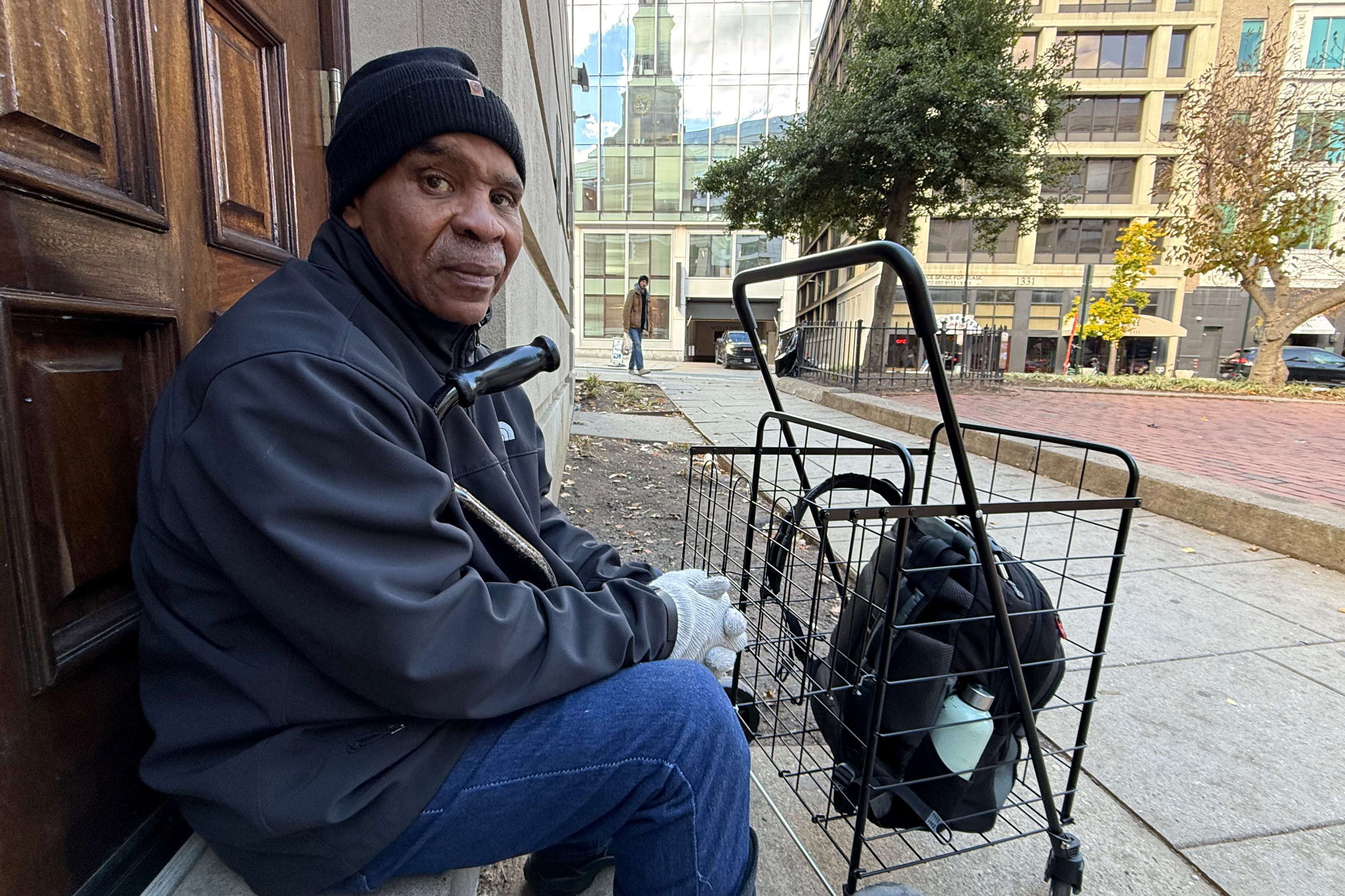 A photo of Chris Jones seated on the steps of a church. Next to him is a small cart with his backpack in it.