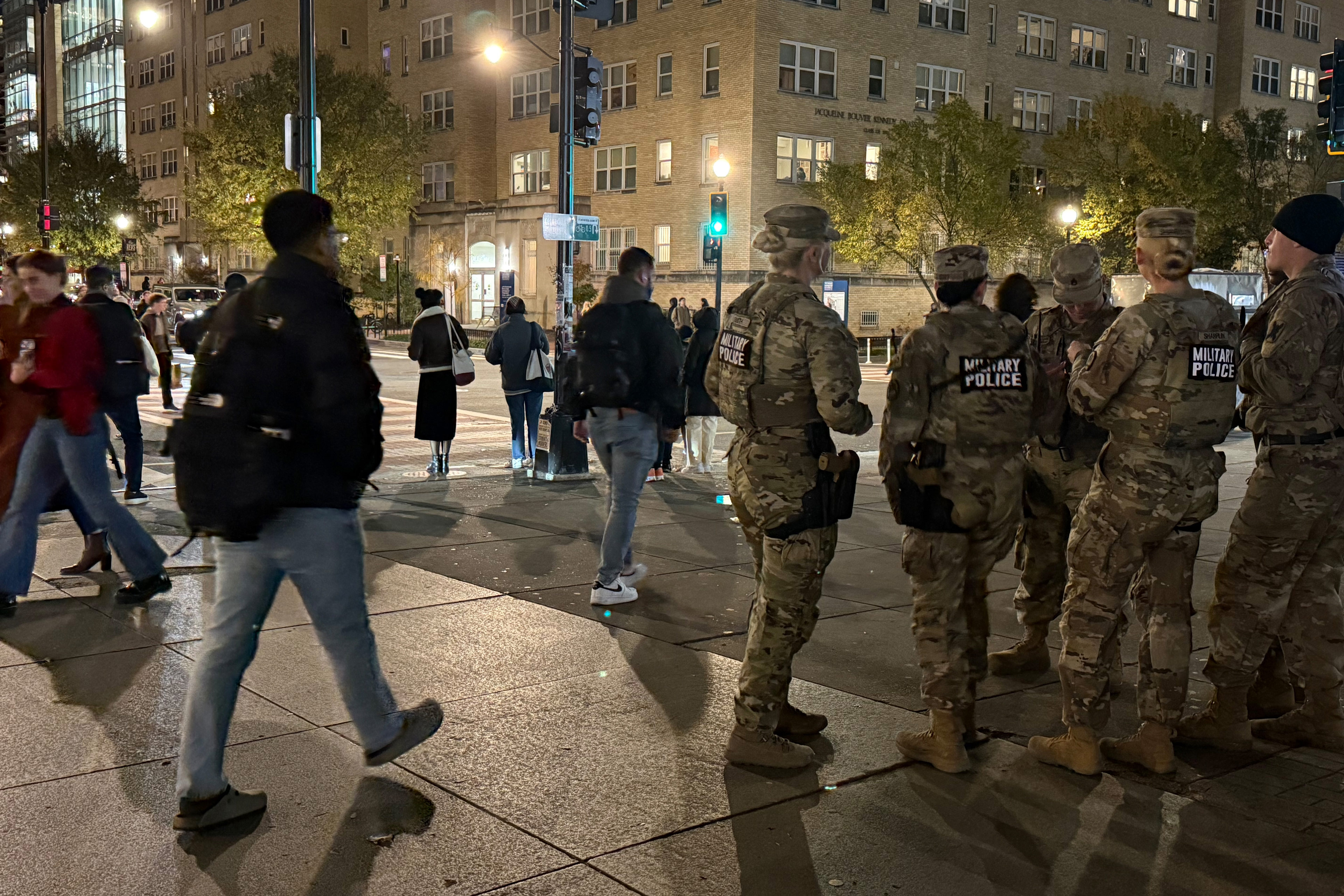 A photo of a D.C. street at night. Pedestrians walk past a set of military police officers in camouflage. 