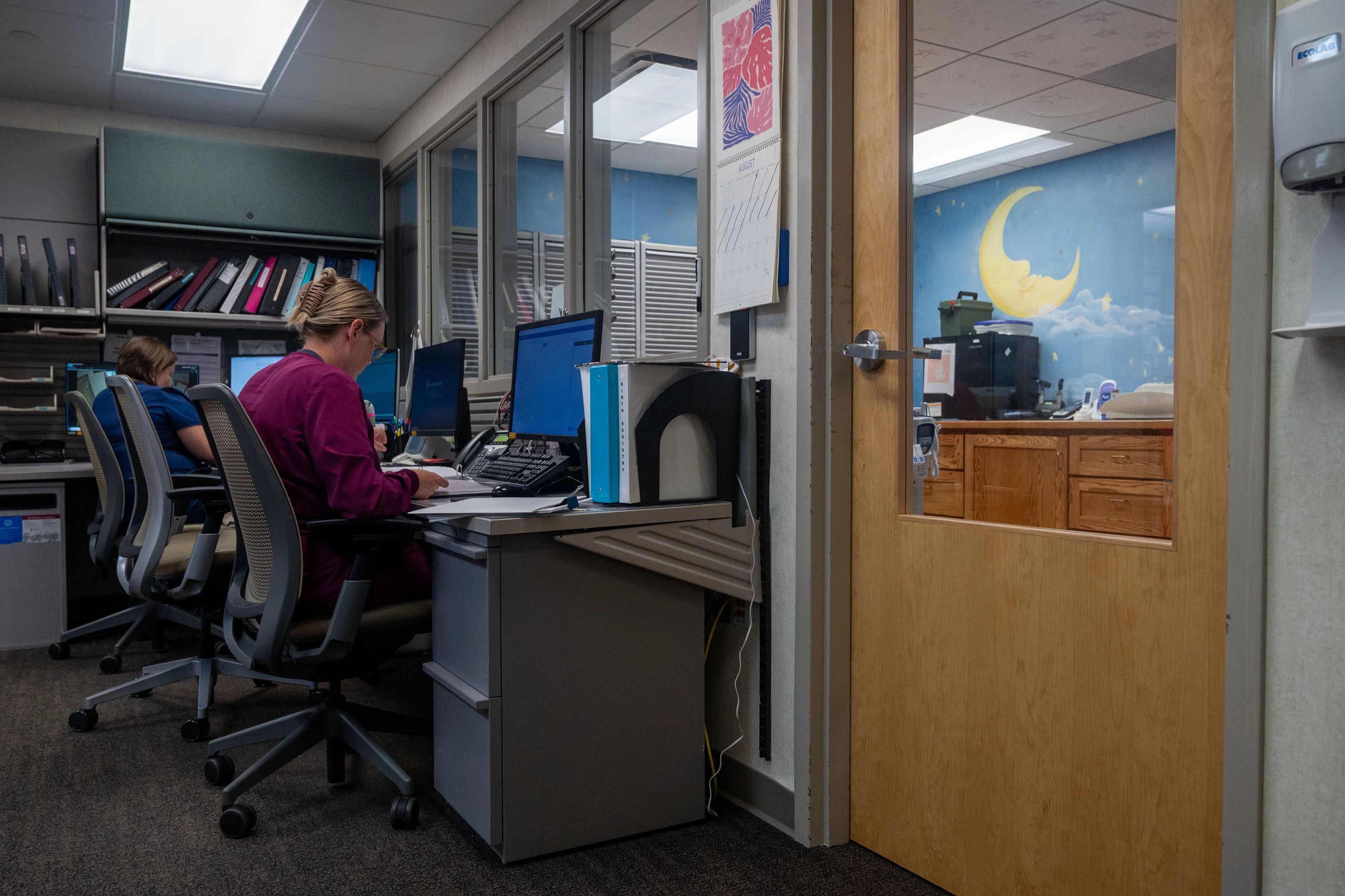 A woman sits at a computer in a small medical reception room.