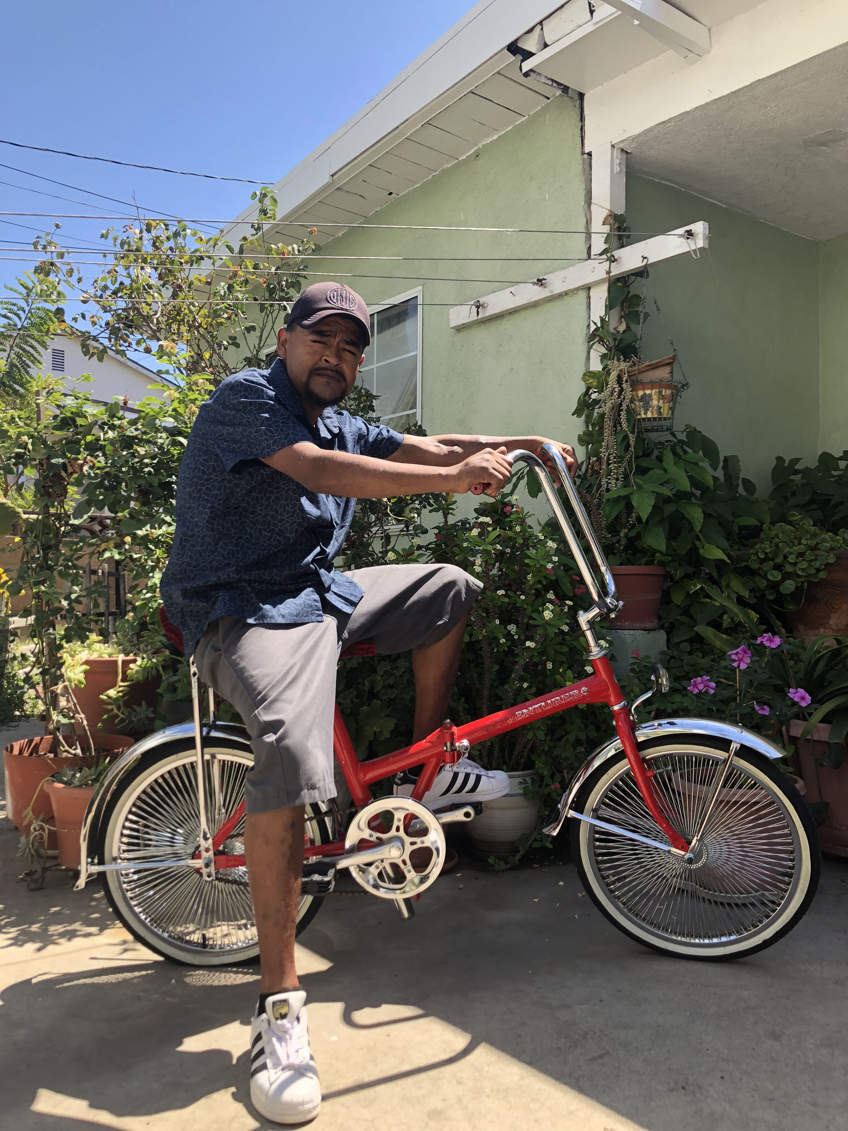 A man sits on his bike in the backyard of his home surrounded by plants and flowers on a sunny day.