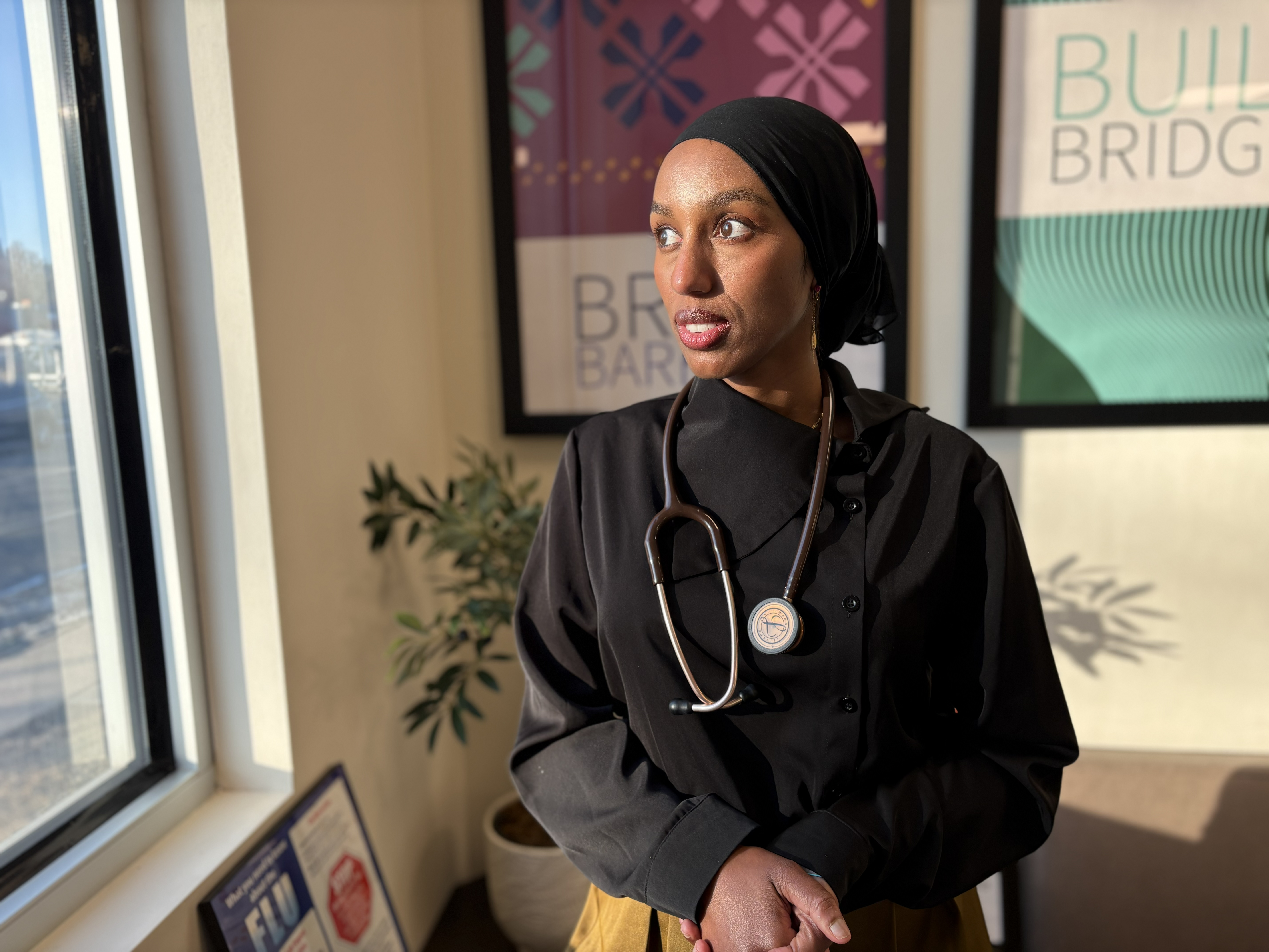 A woman wearing black with her hair covered by a black scarf has a stethoscope draped across her neck. 