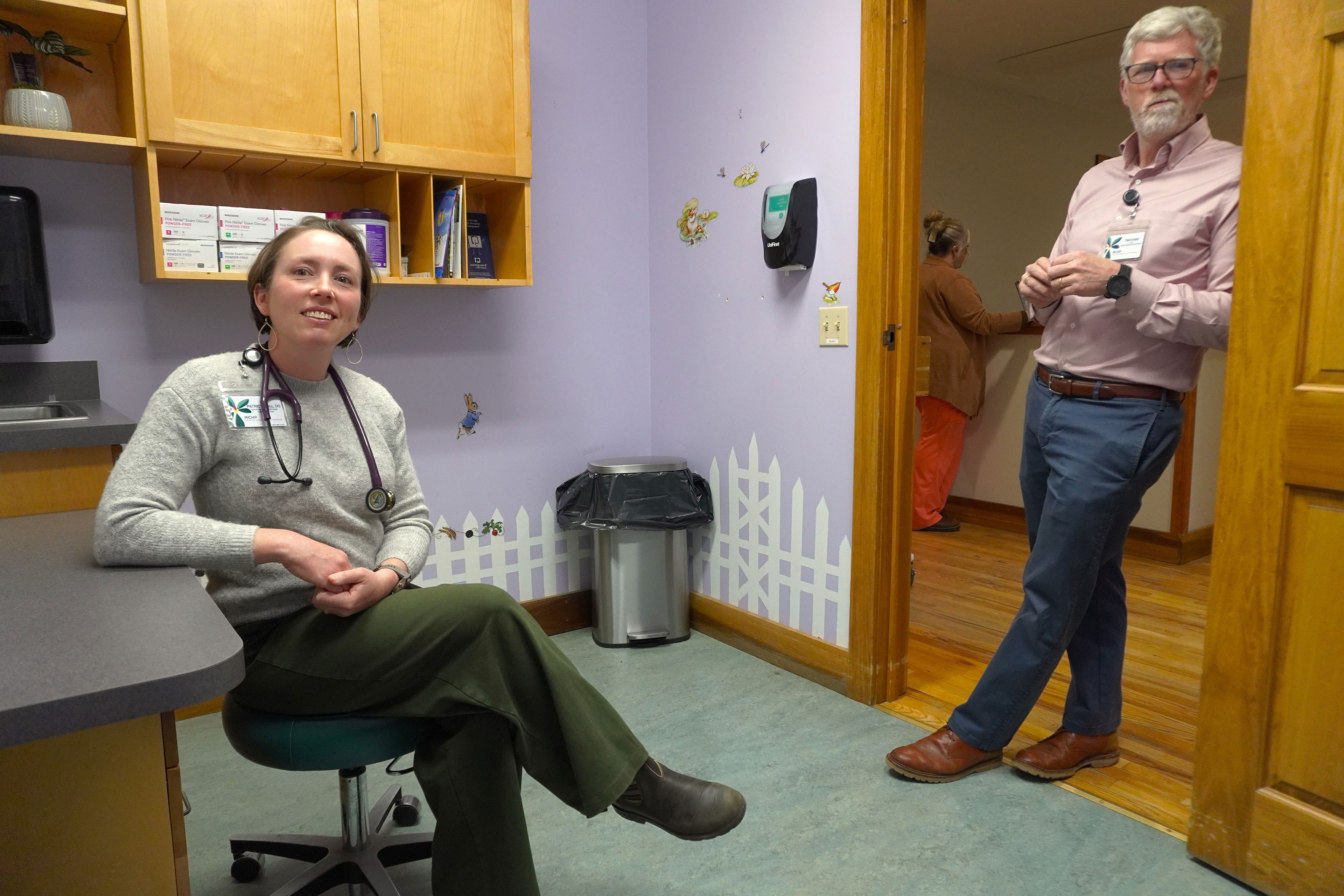 Patricia Hall sits on a stool in Celo Health Center. Tim Evans leans in the doorway.