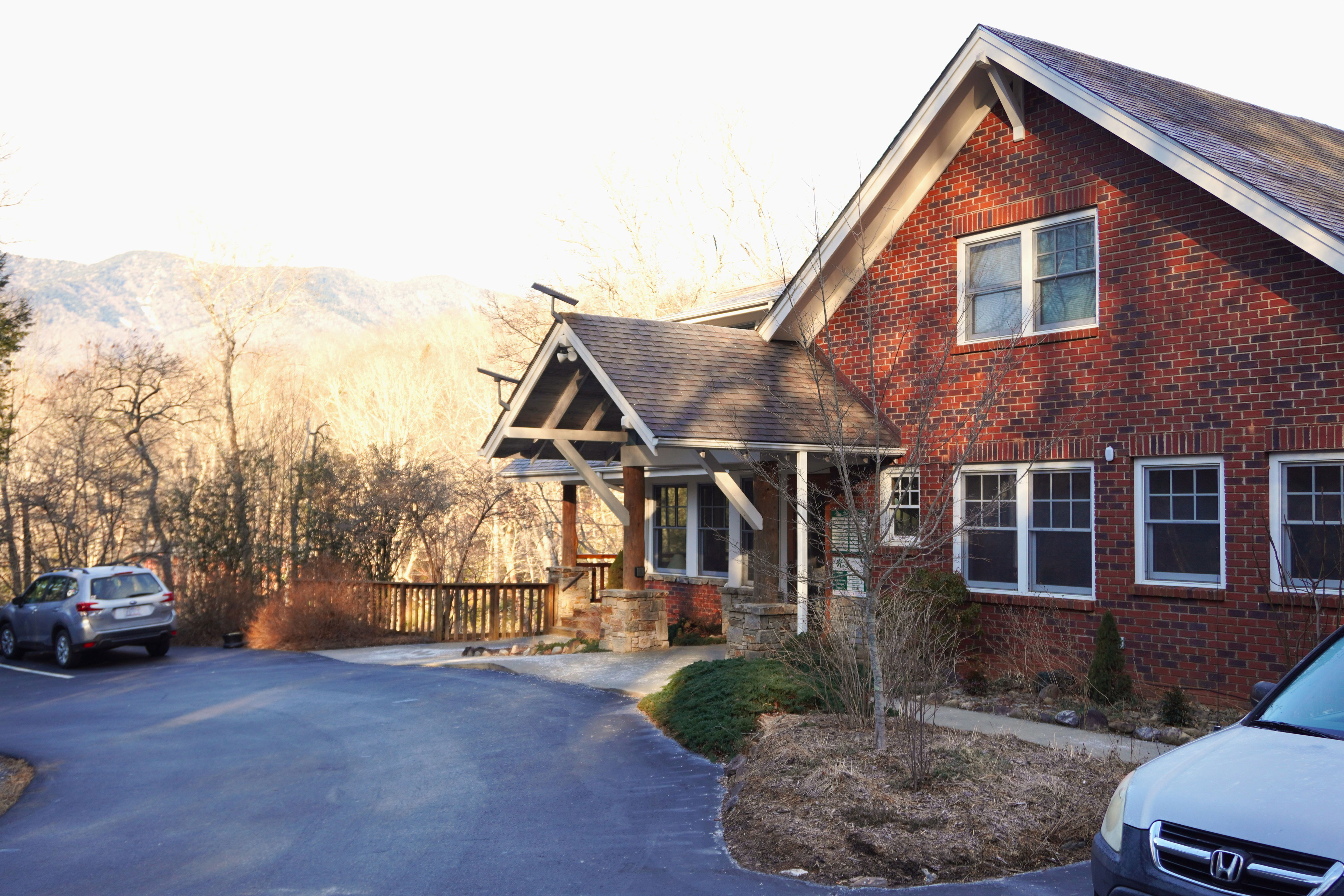 A brick building on a sunny morning with two cars visibly parked in front of it. It is a wooded, mountainous area.